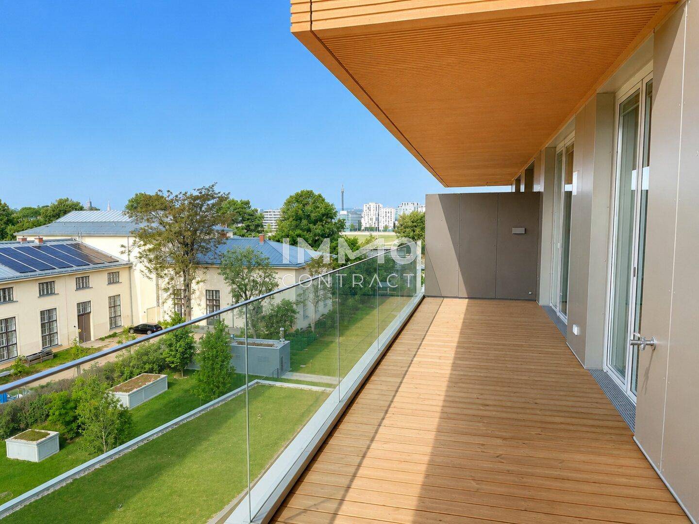 Langer Holzbalkon mit Glasgeländer und Blick auf grüne Anlagen und umliegende Gebäude.