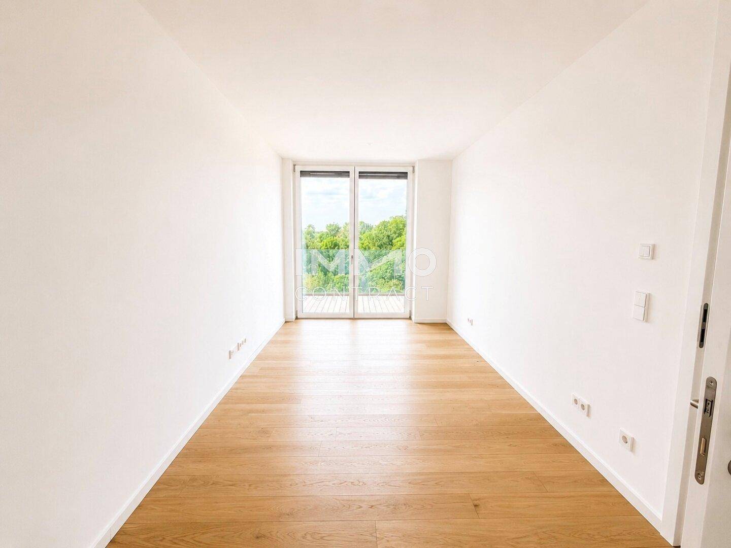 Heller, leerer Raum mit Holzboden und Zugang zu einem Balkon mit grüner Aussicht.