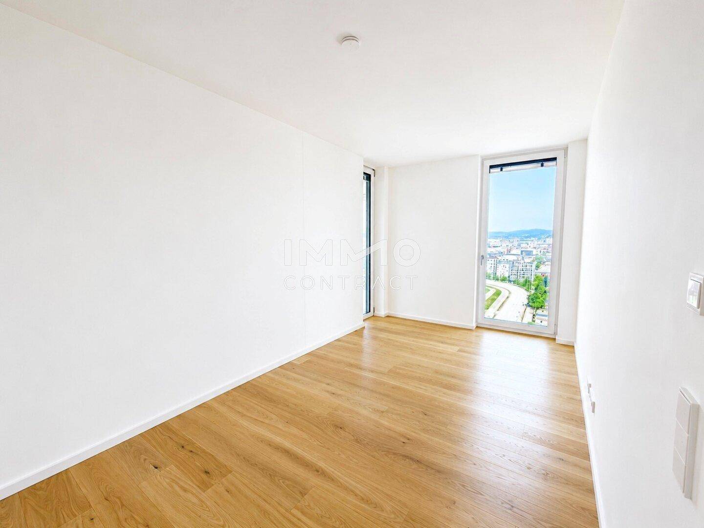 Helles Zimmer mit Holzboden und einem großen Fenster, das einen weiten Blick auf die Stadt bietet.