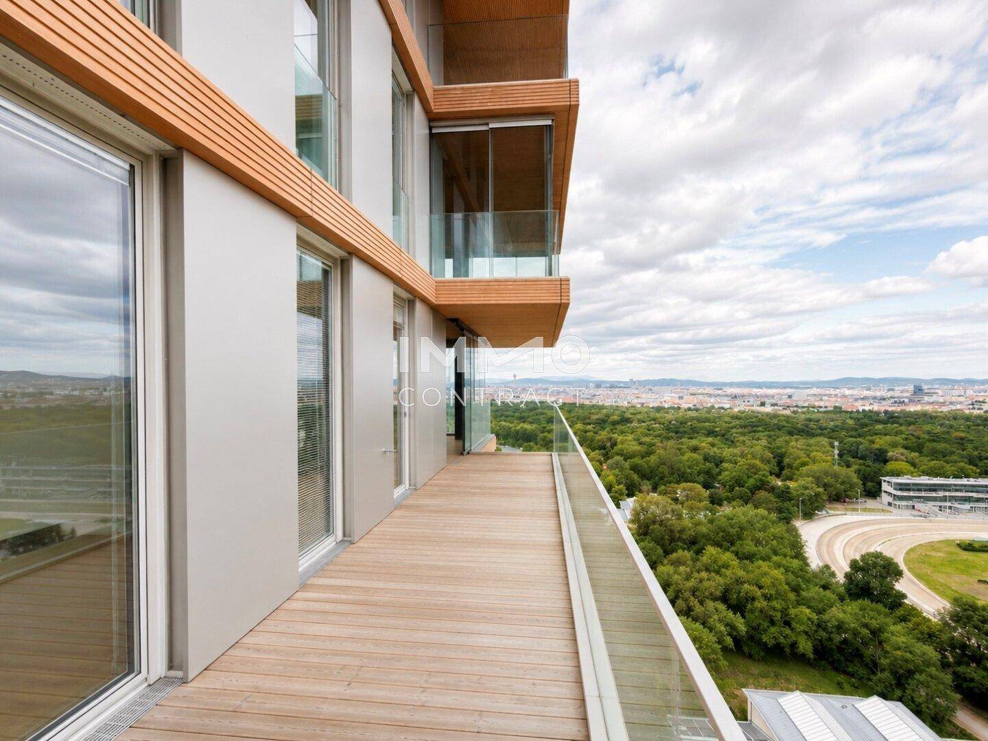 Langer Holzbalkon mit Glasgeländer und weitem Blick über grüne Flächen und die Stadt.