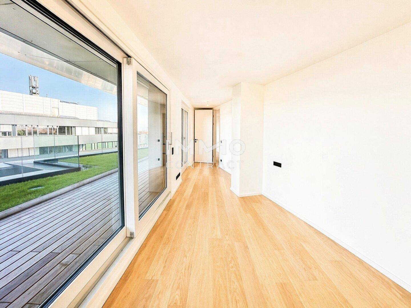 Heller Raum mit Holzparkettboden und großen Fenstern, die auf eine begrünte Dachterrasse blicken.