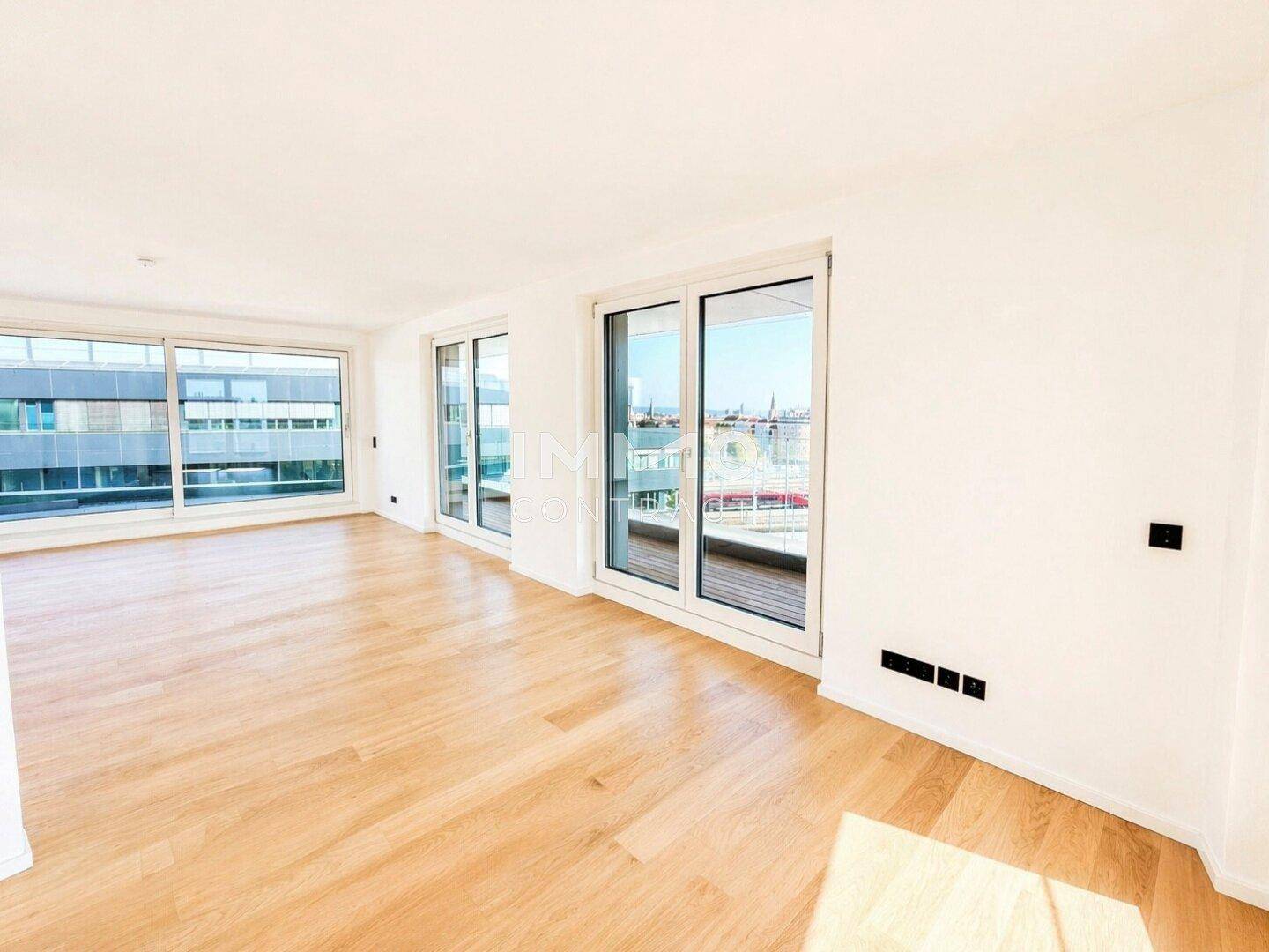 Großes Wohnzimmer mit Holzparkettboden und breiten Fenstern, die viel Tageslicht hereinlassen.