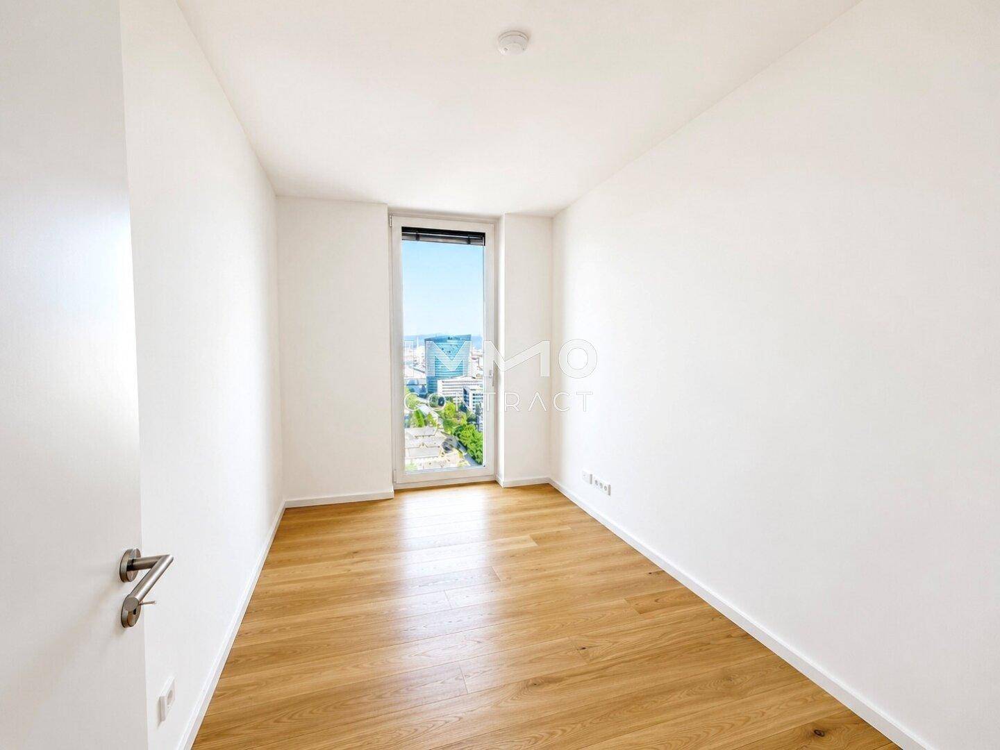 Helles Zimmer mit Holzboden und Fenster, das einen Blick auf die Stadt und die Umgebung ermöglicht.