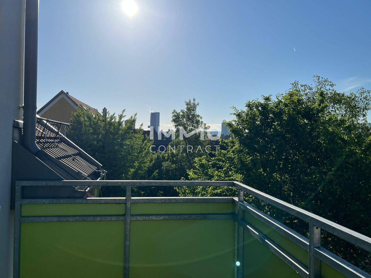 Sonniger Balkon mit grüner Brüstung und Blick auf Bäume sowie die Skyline in der Ferne.