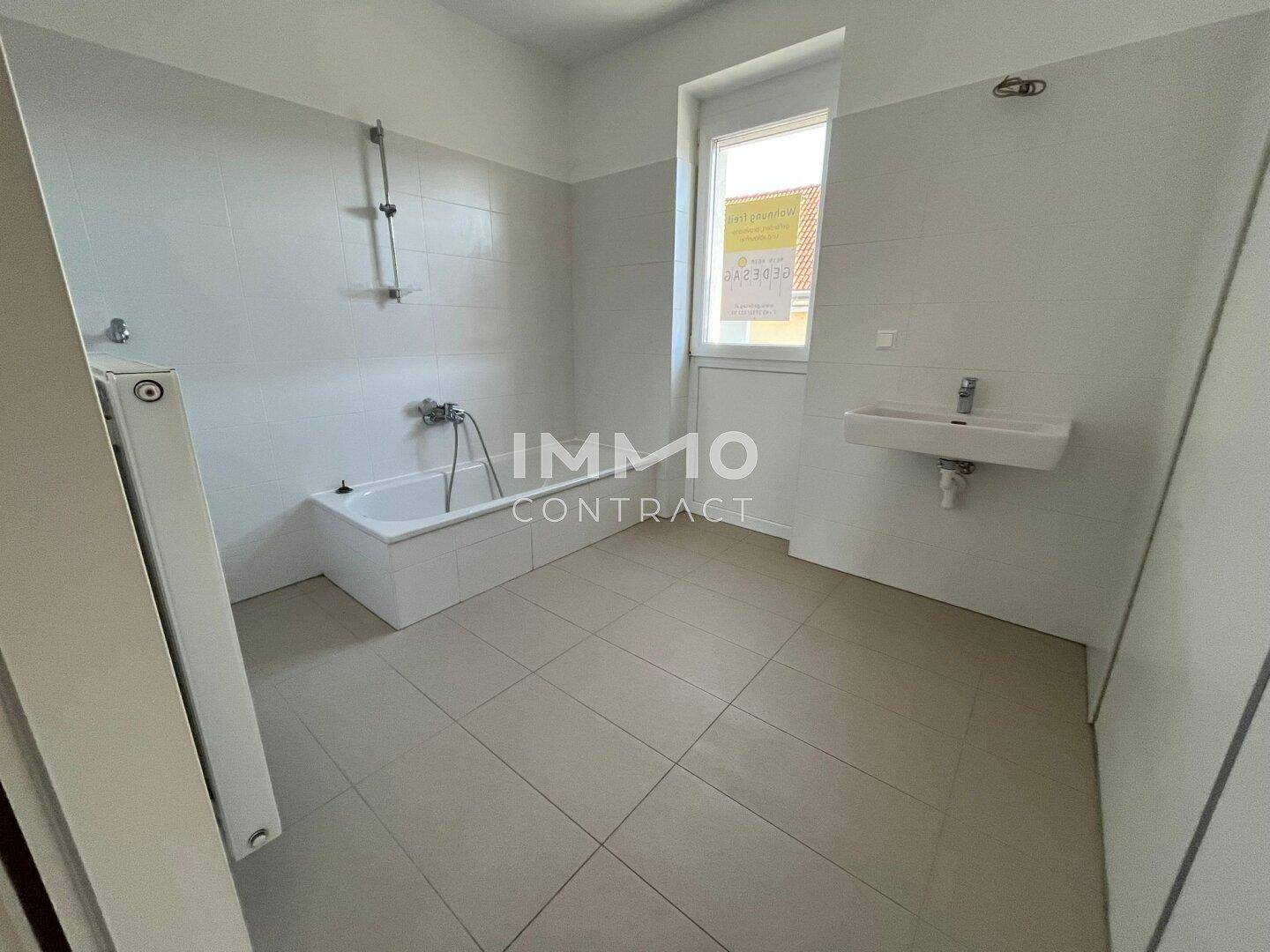 Helles Badezimmer mit Fliesenboden, Badewanne, Waschbecken und Fenster für Tageslicht.
