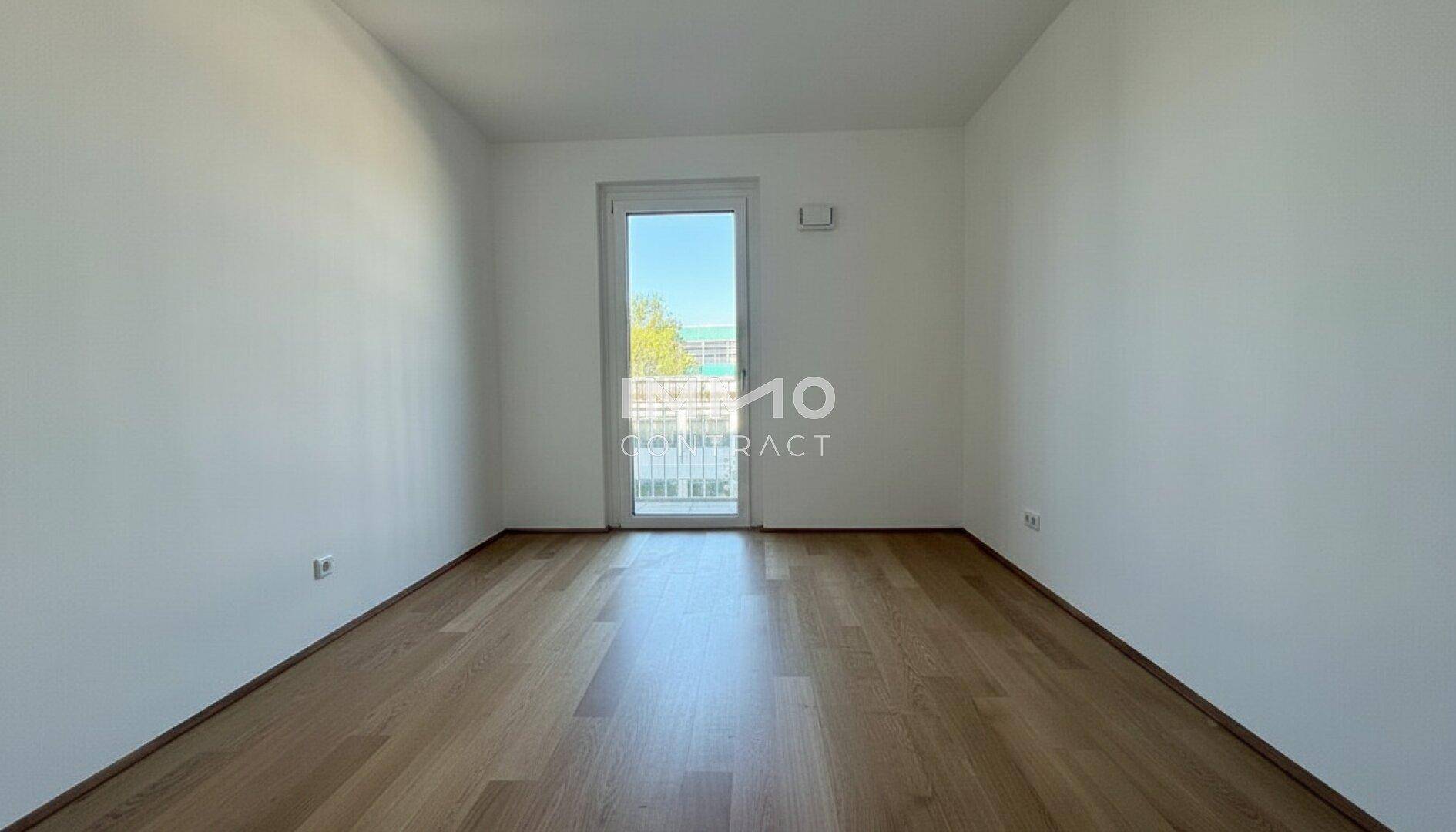 Helles Zimmer mit Parkettboden und Zugang zu einem kleinen Balkon, bietet viel Tageslicht.