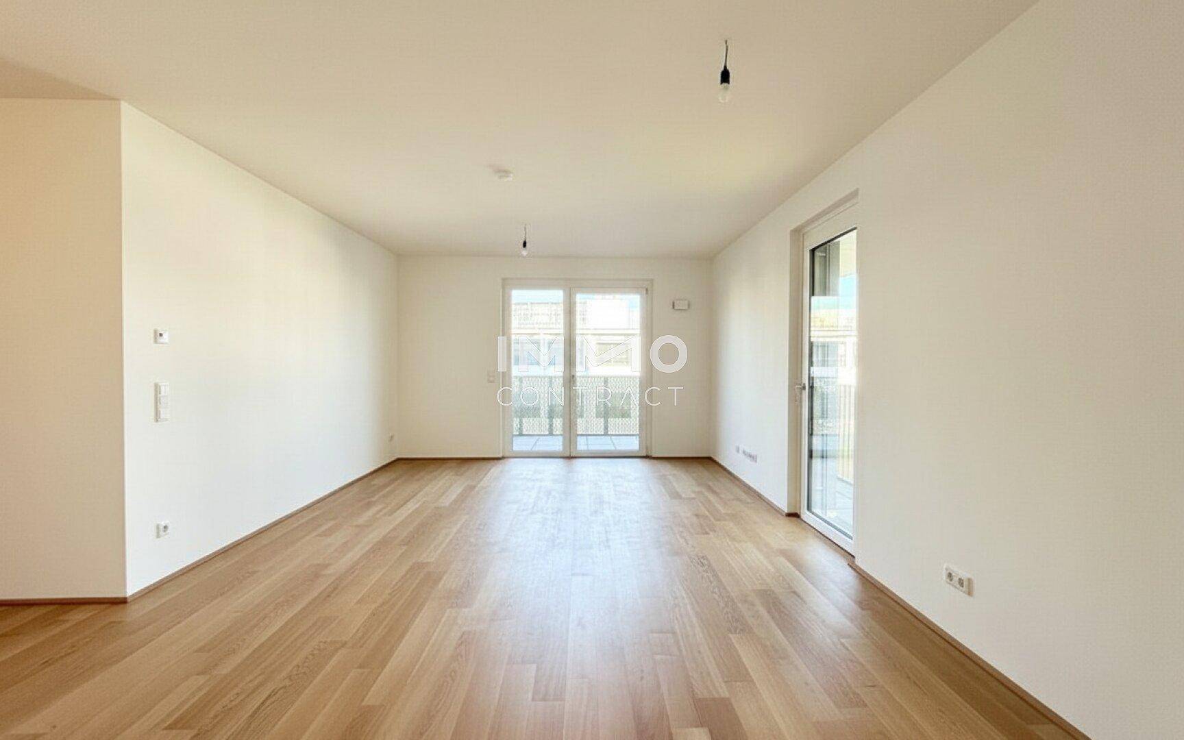 Helles, leeres Wohnzimmer mit Parkettboden und Zugang zu einem Balkon oder einer Terrasse.