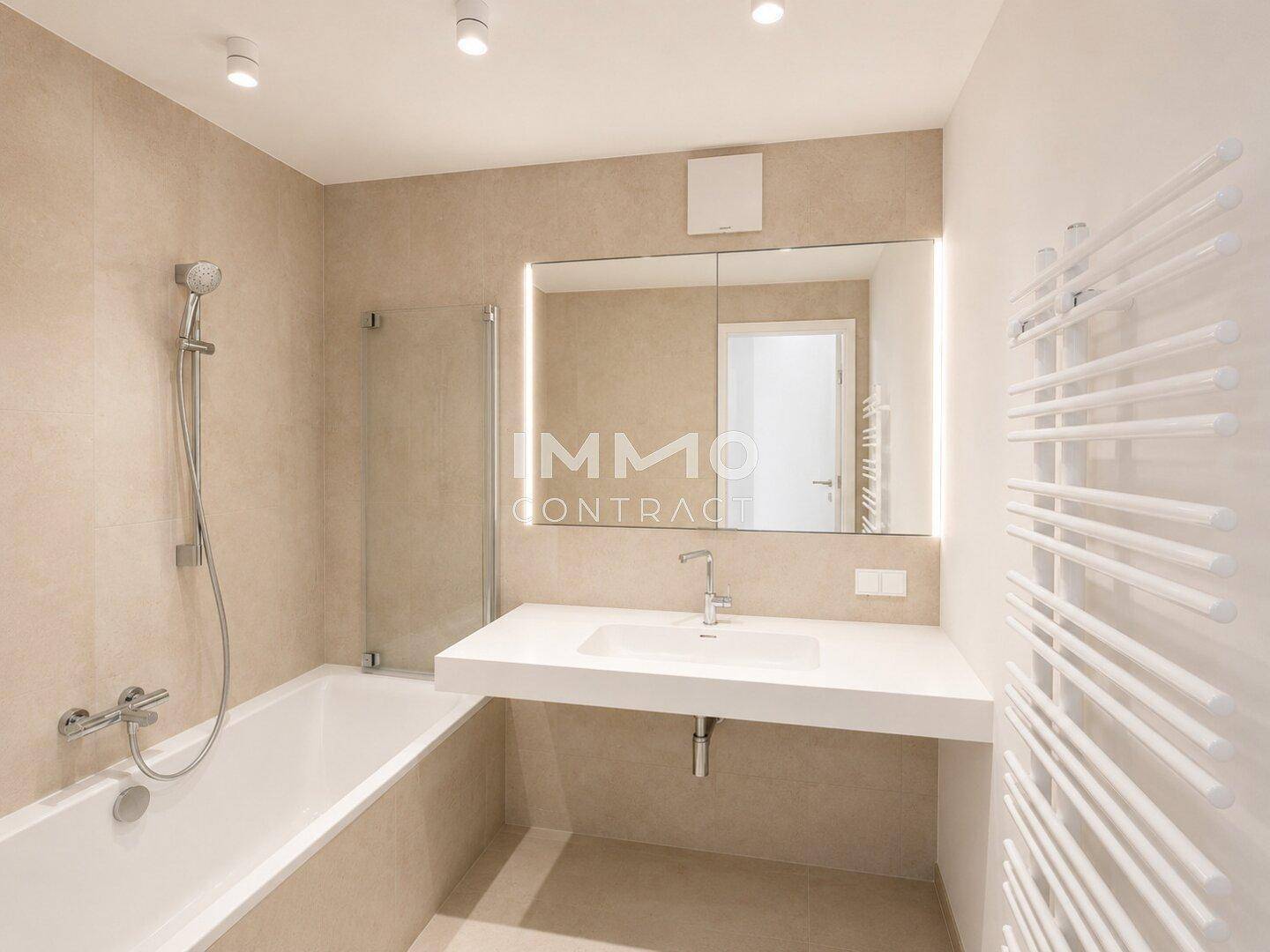 Zeitgemäßes Badezimmer mit Badewanne, beigen Fliesen und einem breiten Waschtisch mit Spiegel.