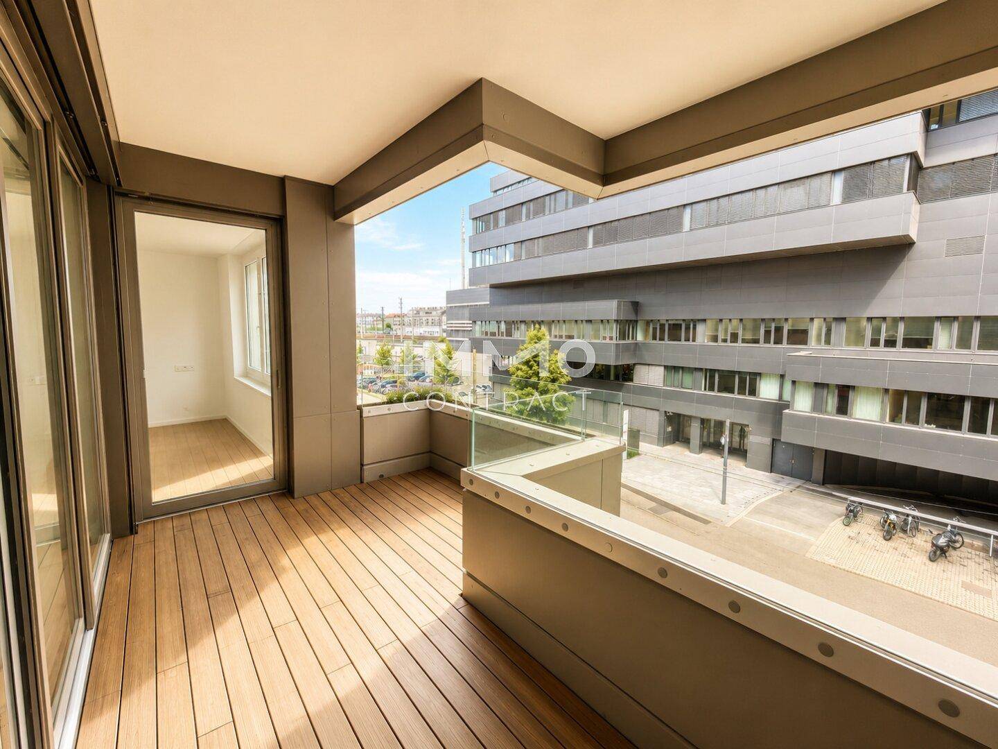 Moderner Balkon mit Holzdeck und Glasgeländer, bietet Blick auf die umliegende Architektur.