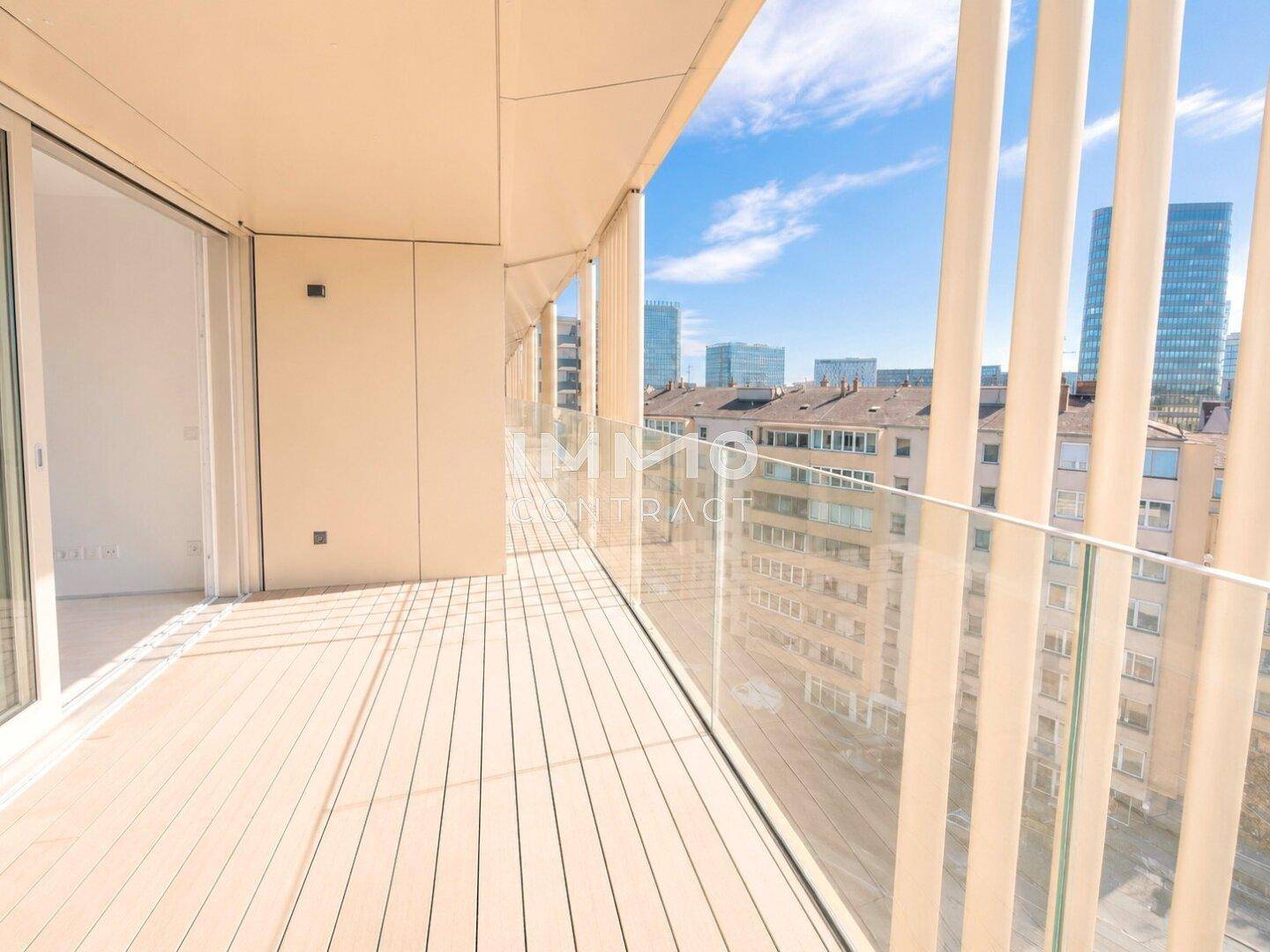 Heller Balkon mit modernem Holzboden und transparentem Glasgeländer, umgeben von Stadtgebäuden.
