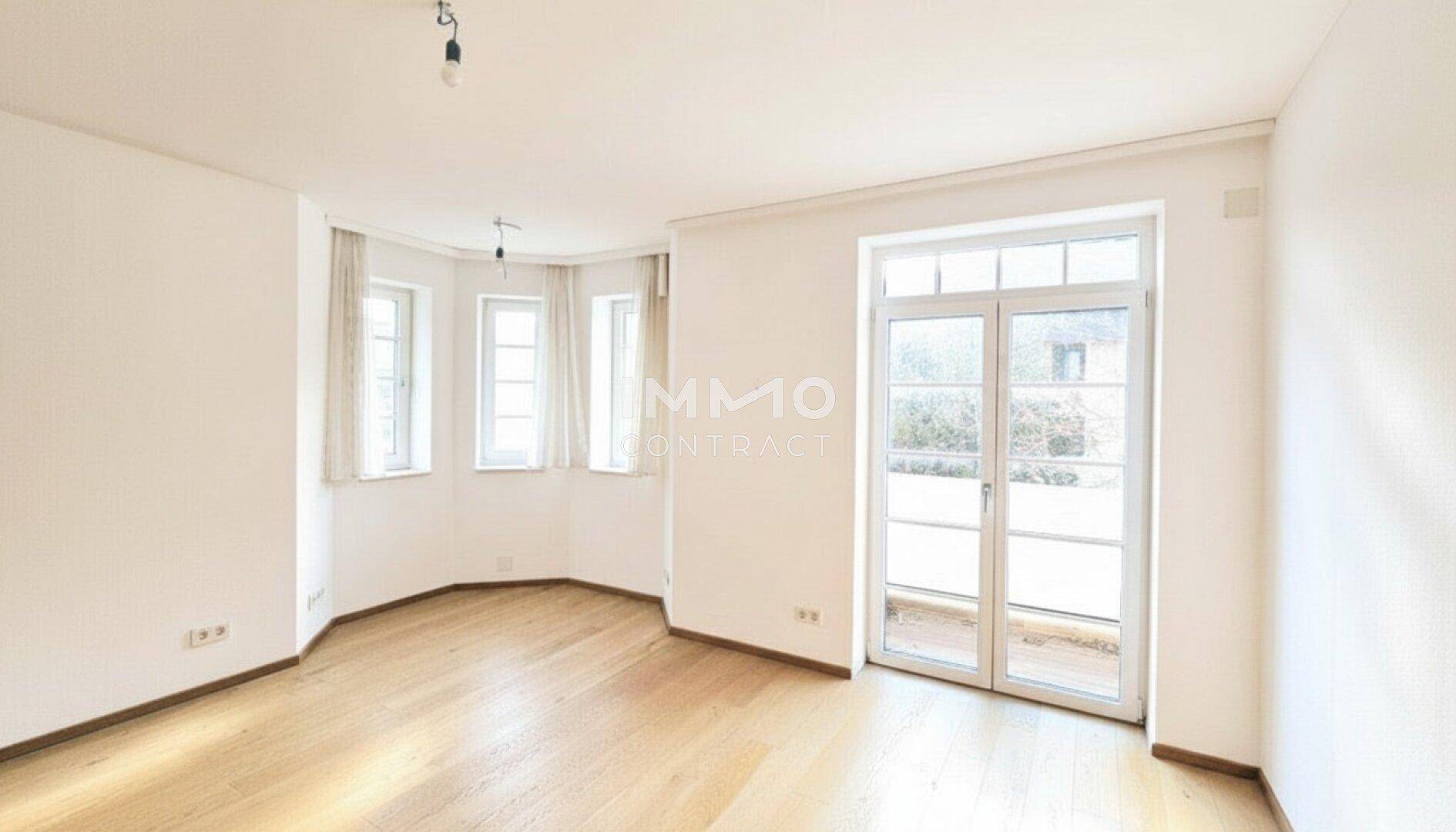 Heller Raum mit Holzboden, Erkerfenstern und Zugang zu einem Balkon, ideal als Wohnzimmer.