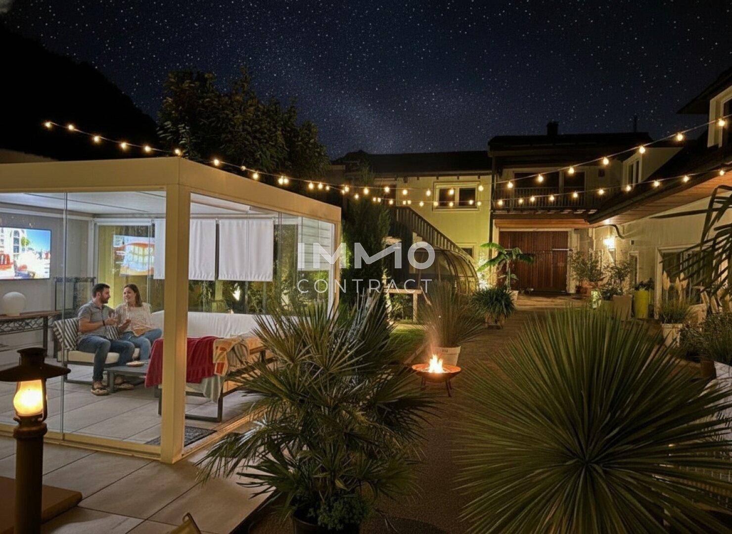 Gemütlicher Außenbereich mit verglaster Terrasse, Lichterketten und Feuerstelle unter sternenklarem Nachthimmel.
