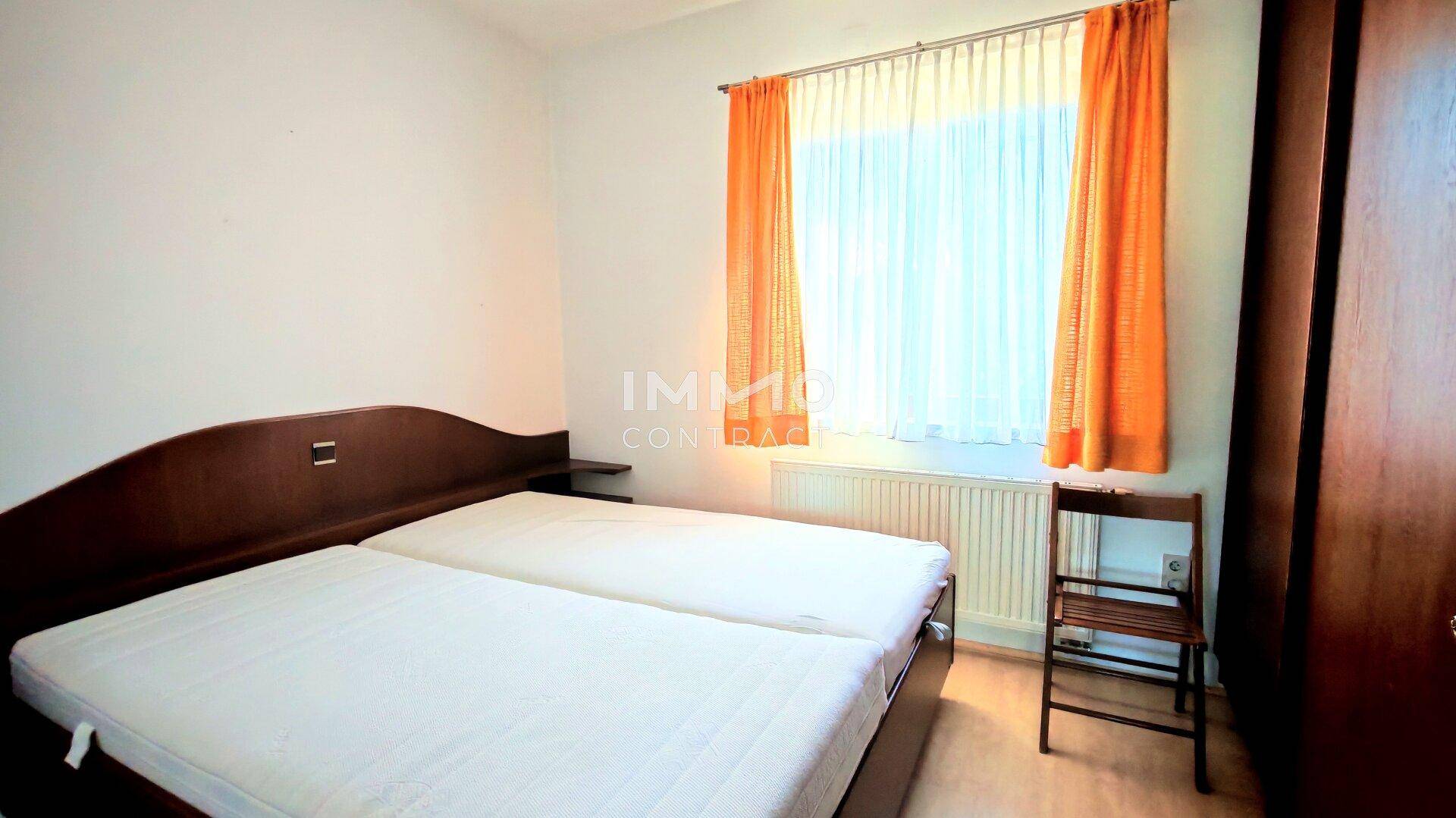 Schlafzimmer mit zwei Einzelbetten, Holzschrank und Fenster mit farbigen Vorhängen.