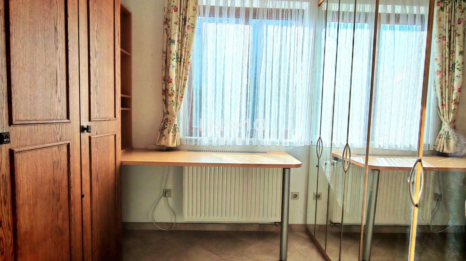 Arbeitsbereich mit Schreibtisch, Holzschrank und Fenstern mit gemusterten Vorhängen.