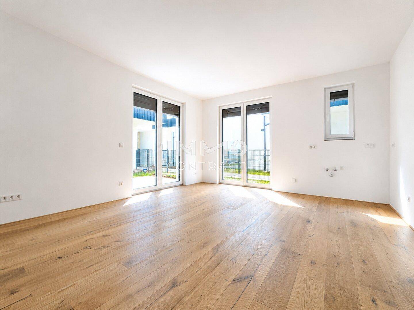 Helles, leeres Wohnzimmer mit großen Fenstern, Holzboden und Zugang zum Außenbereich.