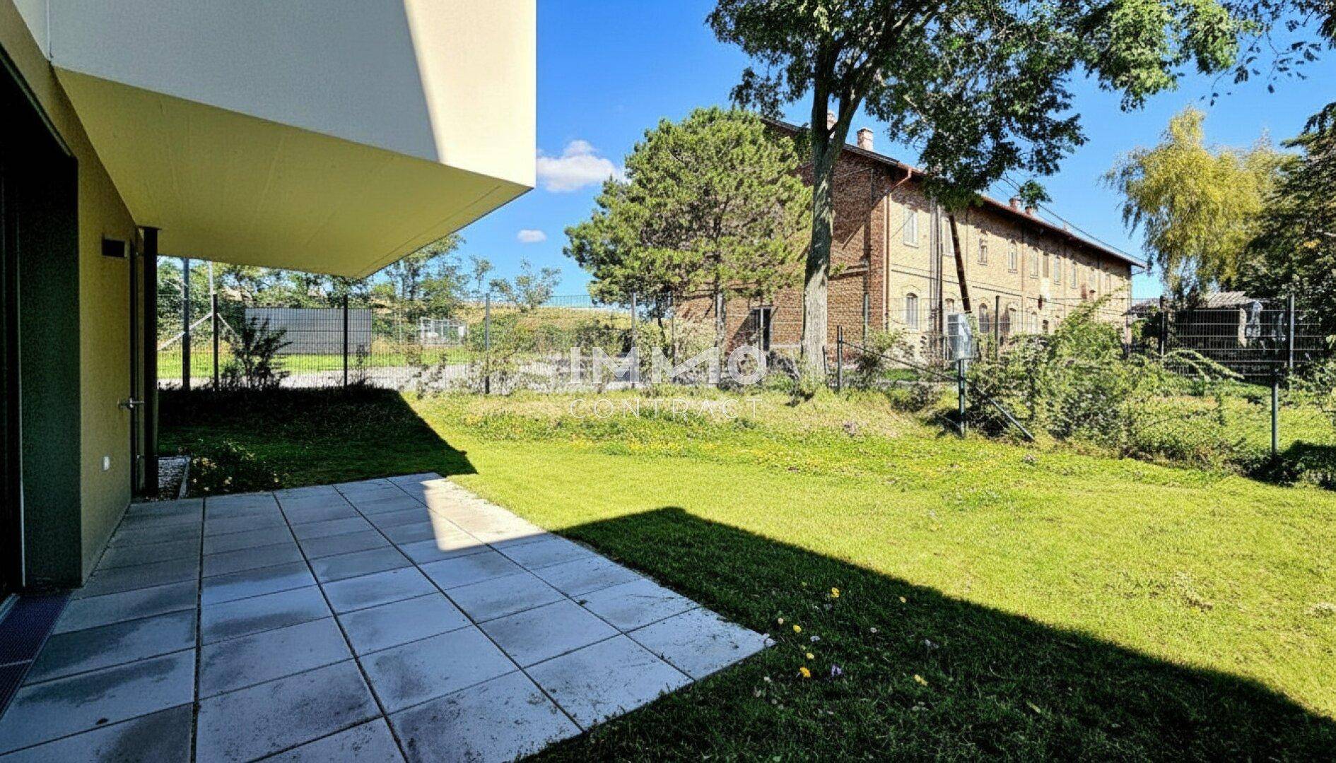 Sonniger Terrassenbereich mit Rasenfläche und Blick auf ein charmantes Backsteingebäude im Hintergrund.