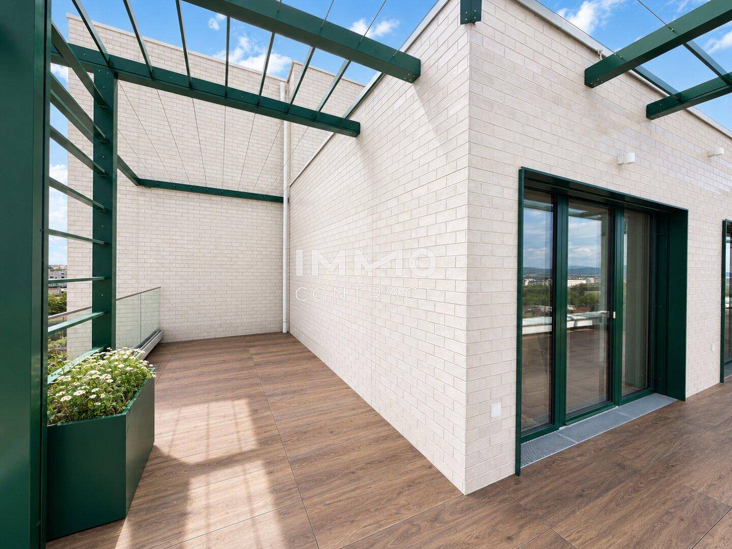 Teil der Terrasse mit Holzfliesen, weißer Ziegelfassade und grüner Pergola-Struktur.