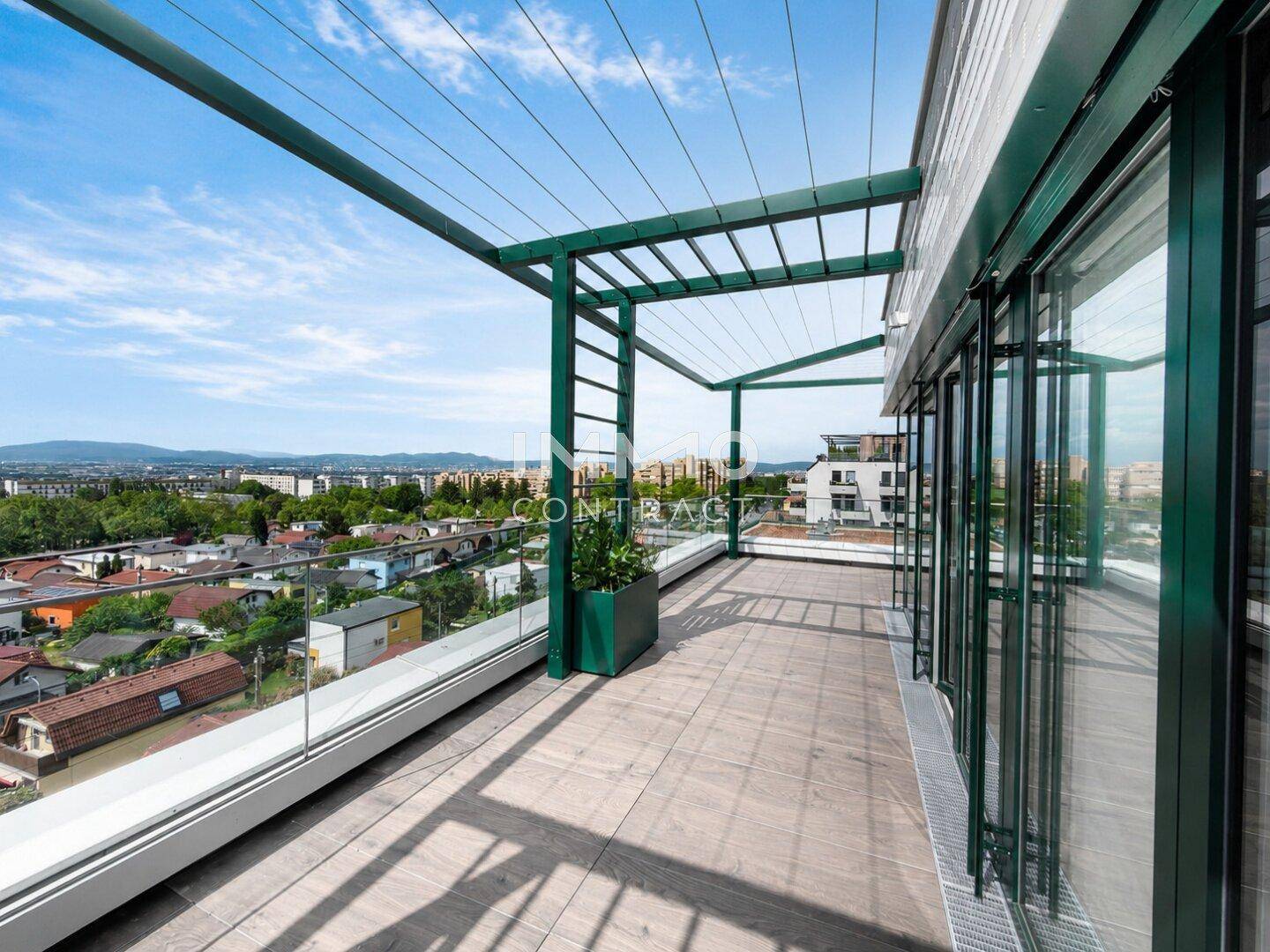 Geräumige Terrasse mit Holzbelag, Glasgeländer und grüner Pergola-Struktur mit Stadtblick.
