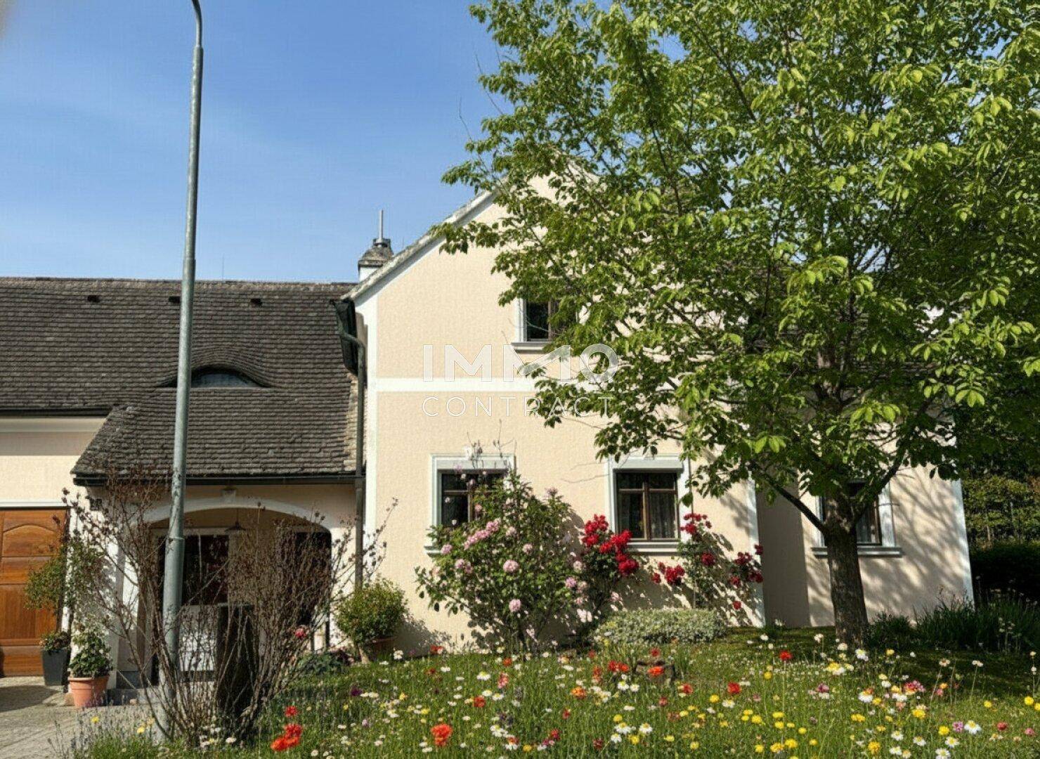 Charmante Hausfassade mit blühenden Rosen und einem großen Baum im Vorgarten unter blauem Himmel.