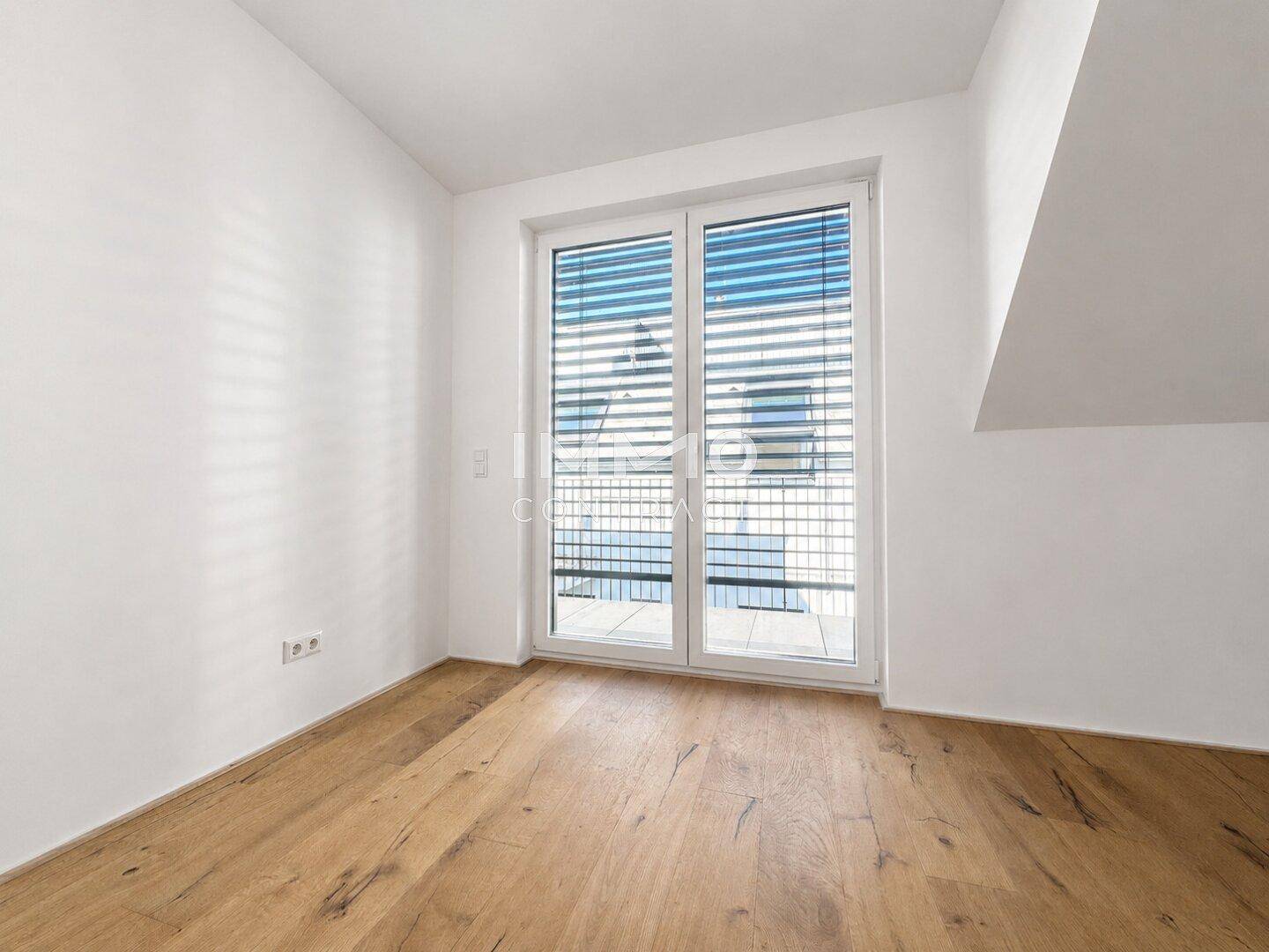 Helles Schlafzimmer mit Holzboden, Dachschräge und Zugang zu einem Balkon durch eine große Glastür.