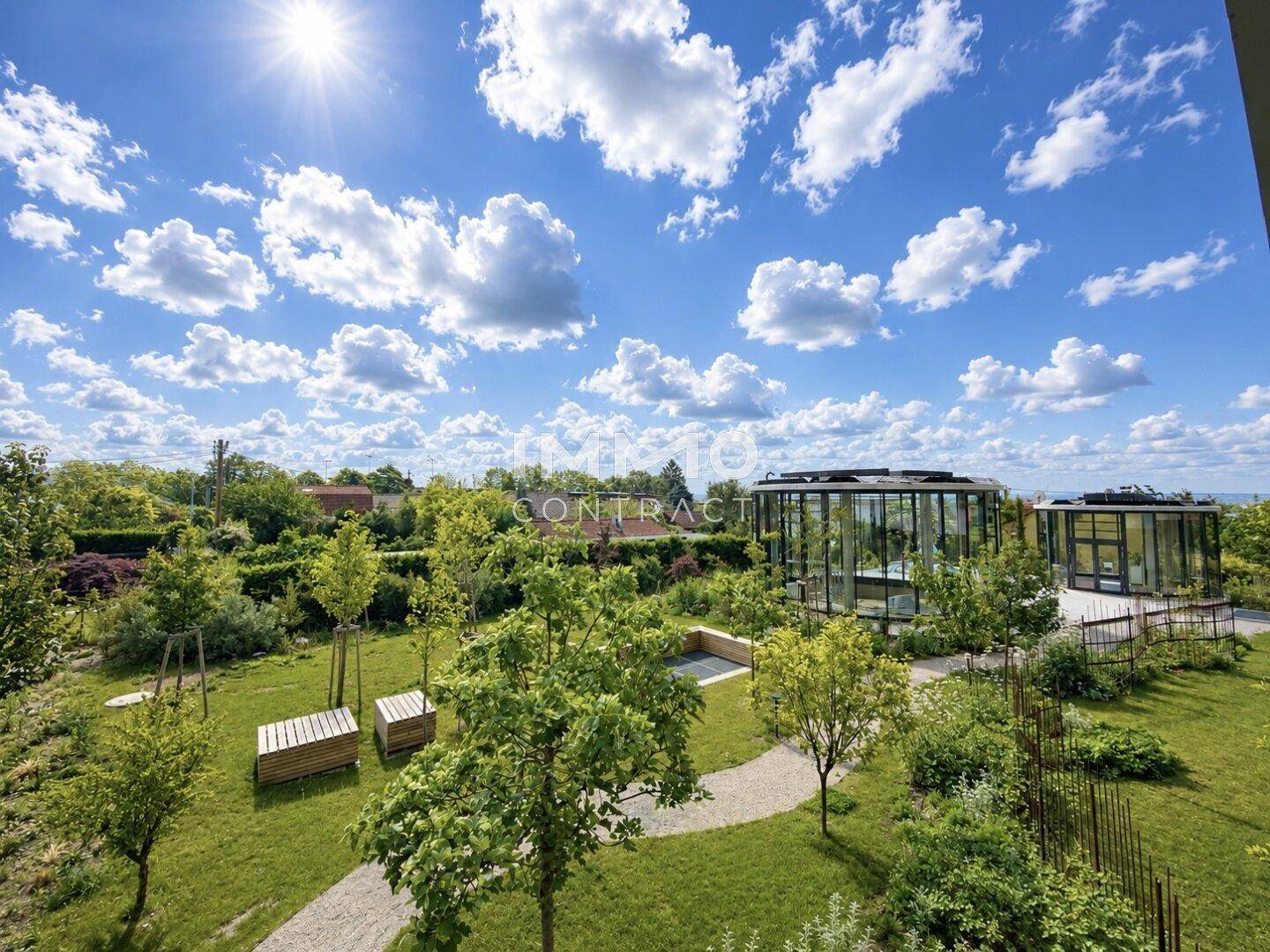 Gepflegte Gartenanlage mit Bäumen, Wegen und modernen Glaspavillons unter blauem Himmel.