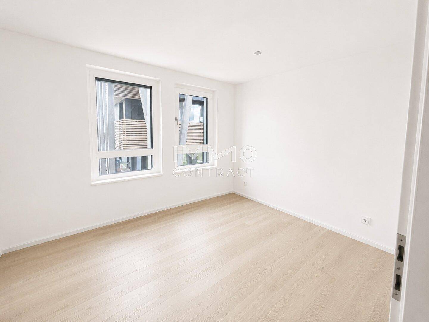 Geräumiges Zimmer mit hellem Holzboden und zwei Fenstern, ideal als Schlaf- oder Arbeitszimmer.