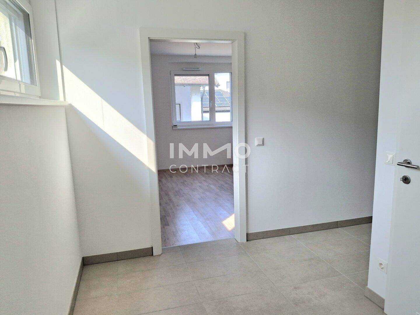 Heller Flur mit Fliesenboden, der den Blick in ein weiteres Zimmer mit Laminatboden freigibt.