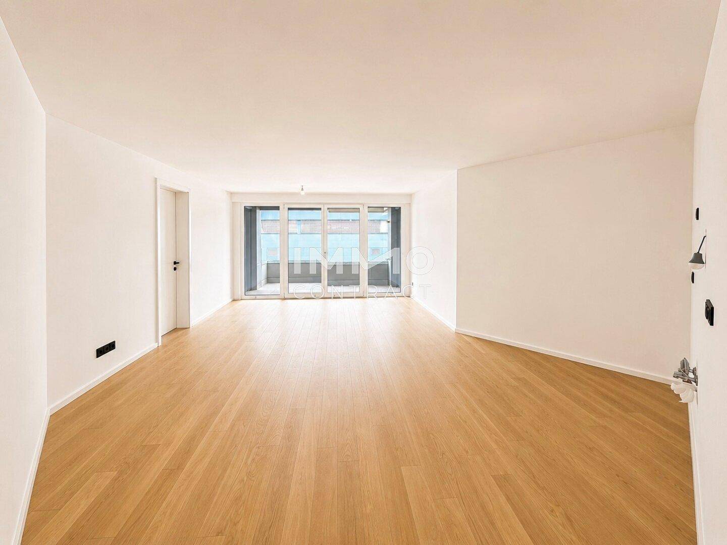 Offener Wohnbereich mit hellem Parkettboden und breiter Fensterfront zum Balkon.