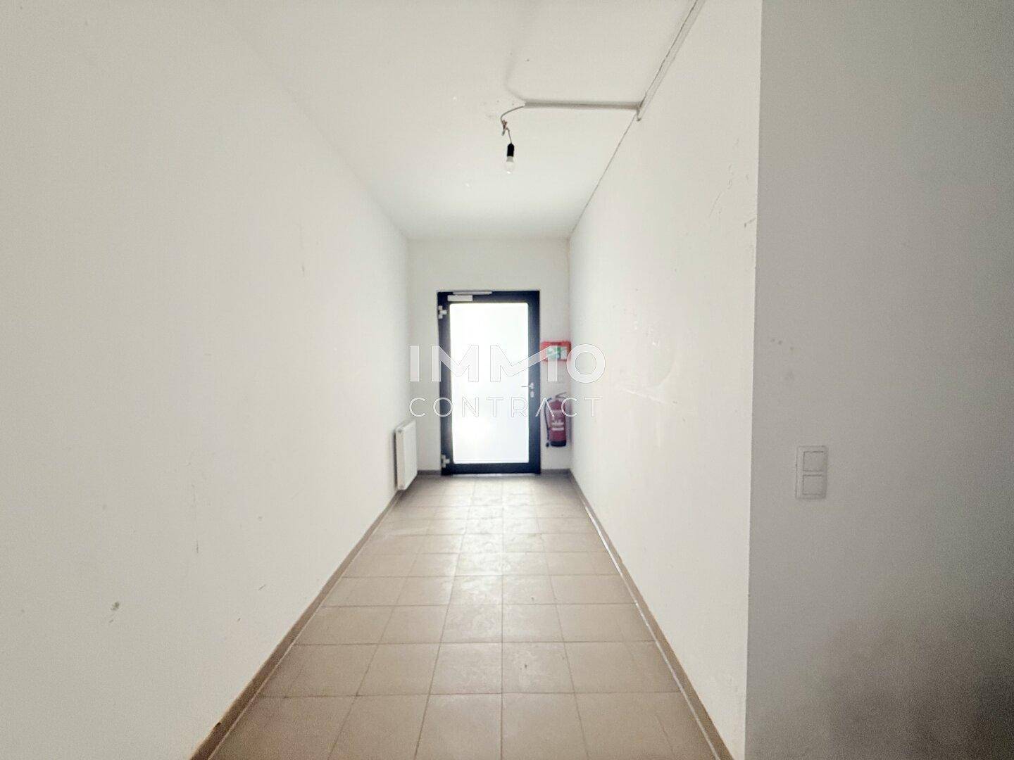 Langer, schmaler Flur mit Fliesenboden, der zum Ausgang und einem Feuerlöscher führt.