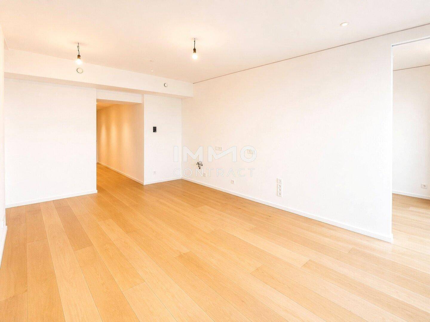 Geräumiges, leeres Zimmer mit hellem Holzboden und schlichten weißen Wänden.