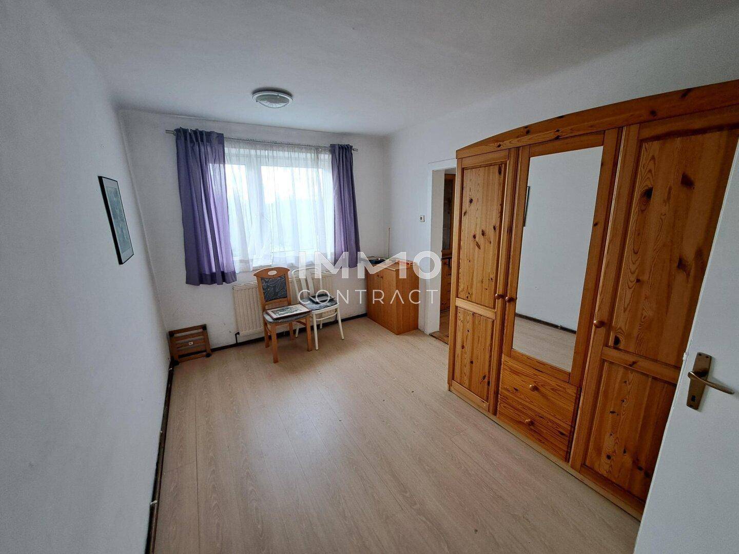 Schlafzimmer mit Laminatboden, großem Holzschrank und Fenster mit lila Vorhängen.
