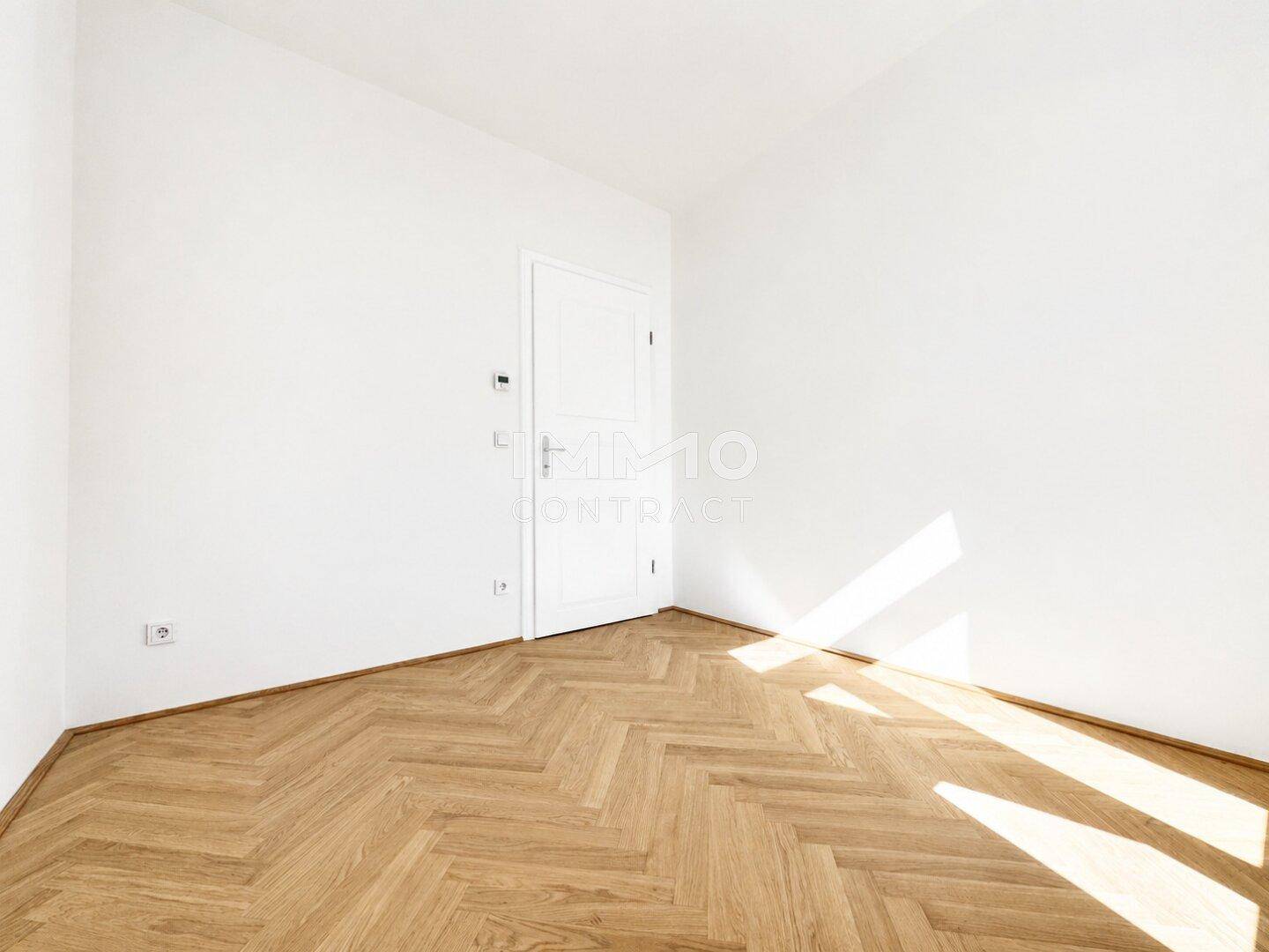 Leeres Zimmer mit hellem Fischgrätparkett und viel natürlichem Lichteinfall durch das Fenster.