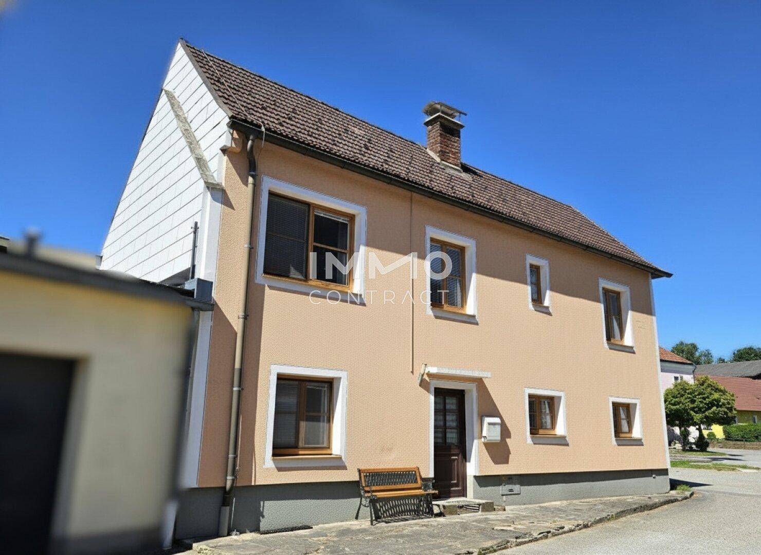 Seitenansicht des Hauses mit beiger Fassade, Satteldach und Holzelementen unter klarem Himmel.