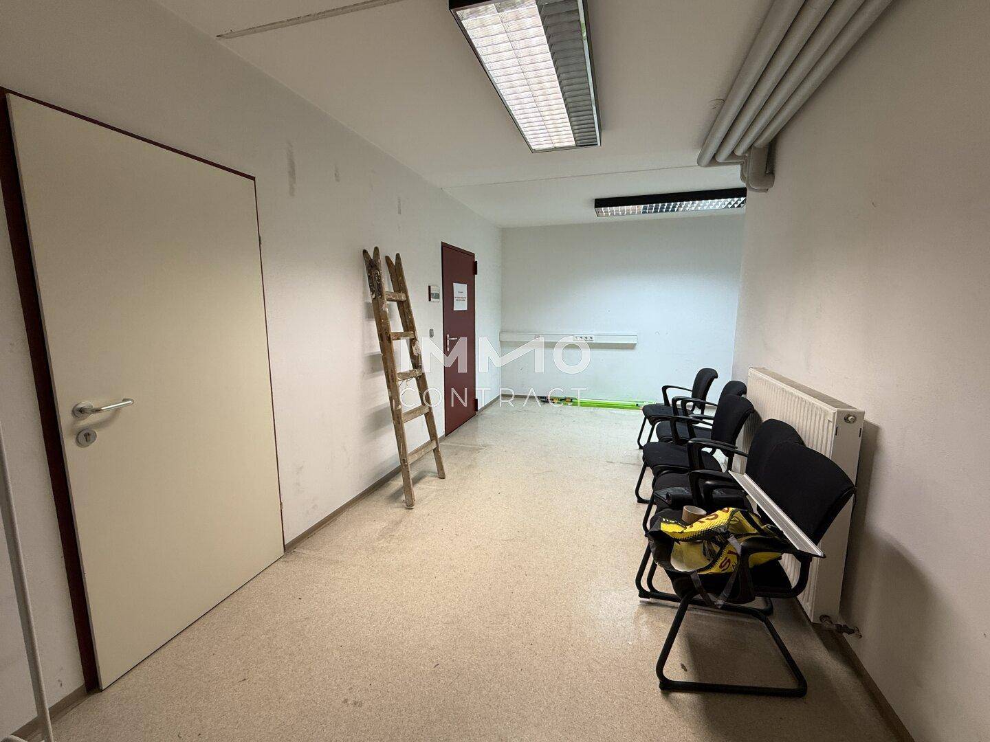 Langer Flur mit Bürostühlen, Heizkörper und Türen zu weiteren Räumen.