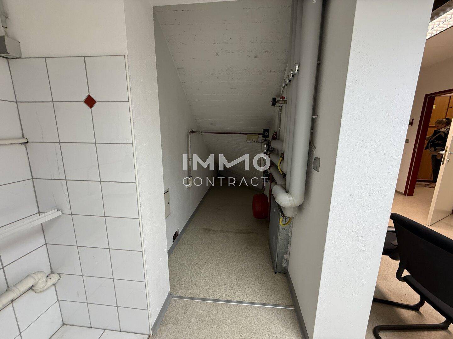 Schmaler Flur mit Rohren an der Decke und Zugang zu einem weiteren Raum.