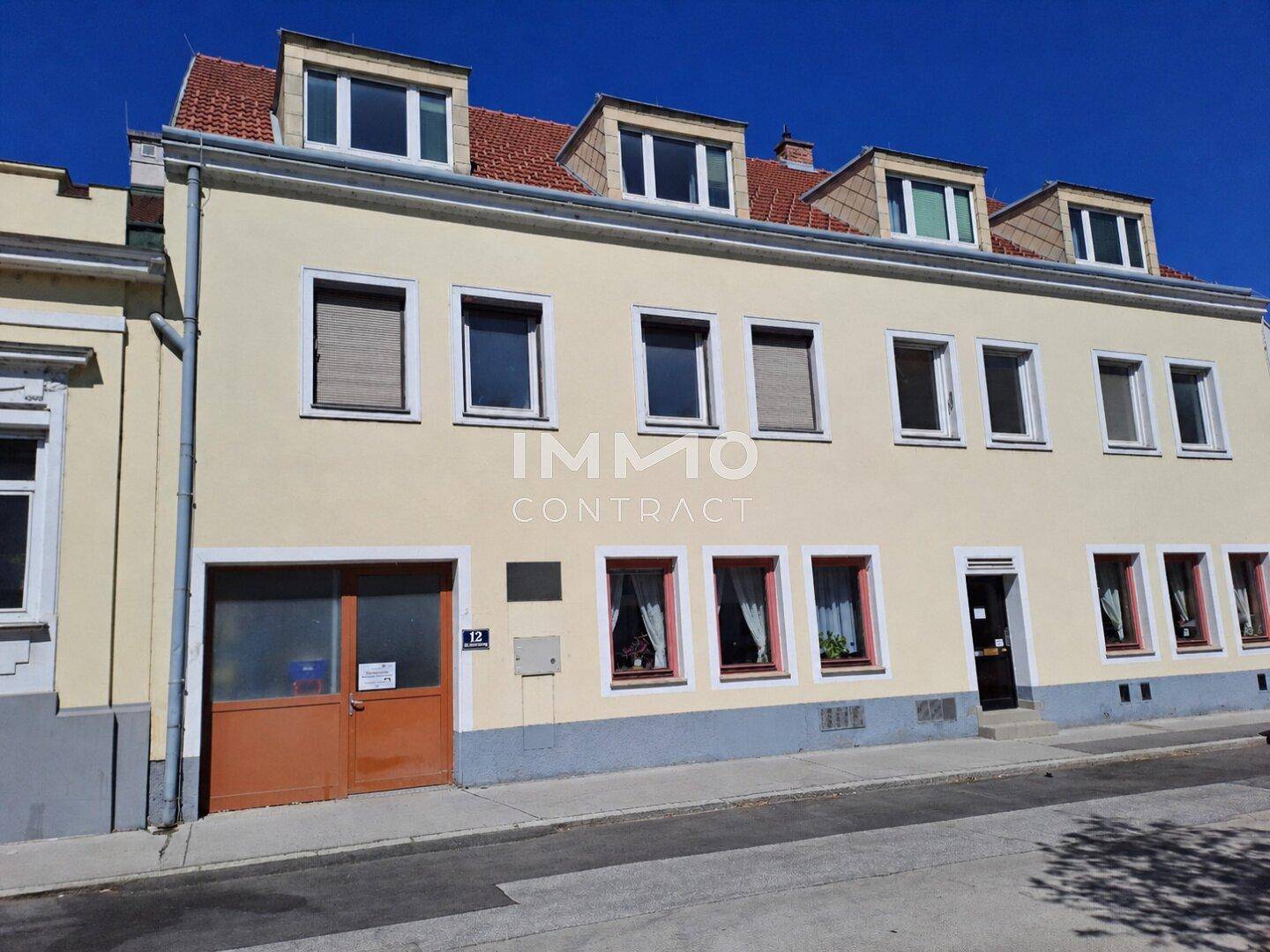 Mehrstöckiges Gebäude mit gelber Fassade, Dachgauben und einer Garageneinfahrt an der Straße.