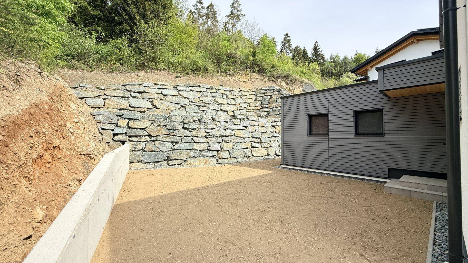 Unfertiger Außenbereich neben dem Haus mit großer Natursteinmauer und vorbereitetem Untergrund.