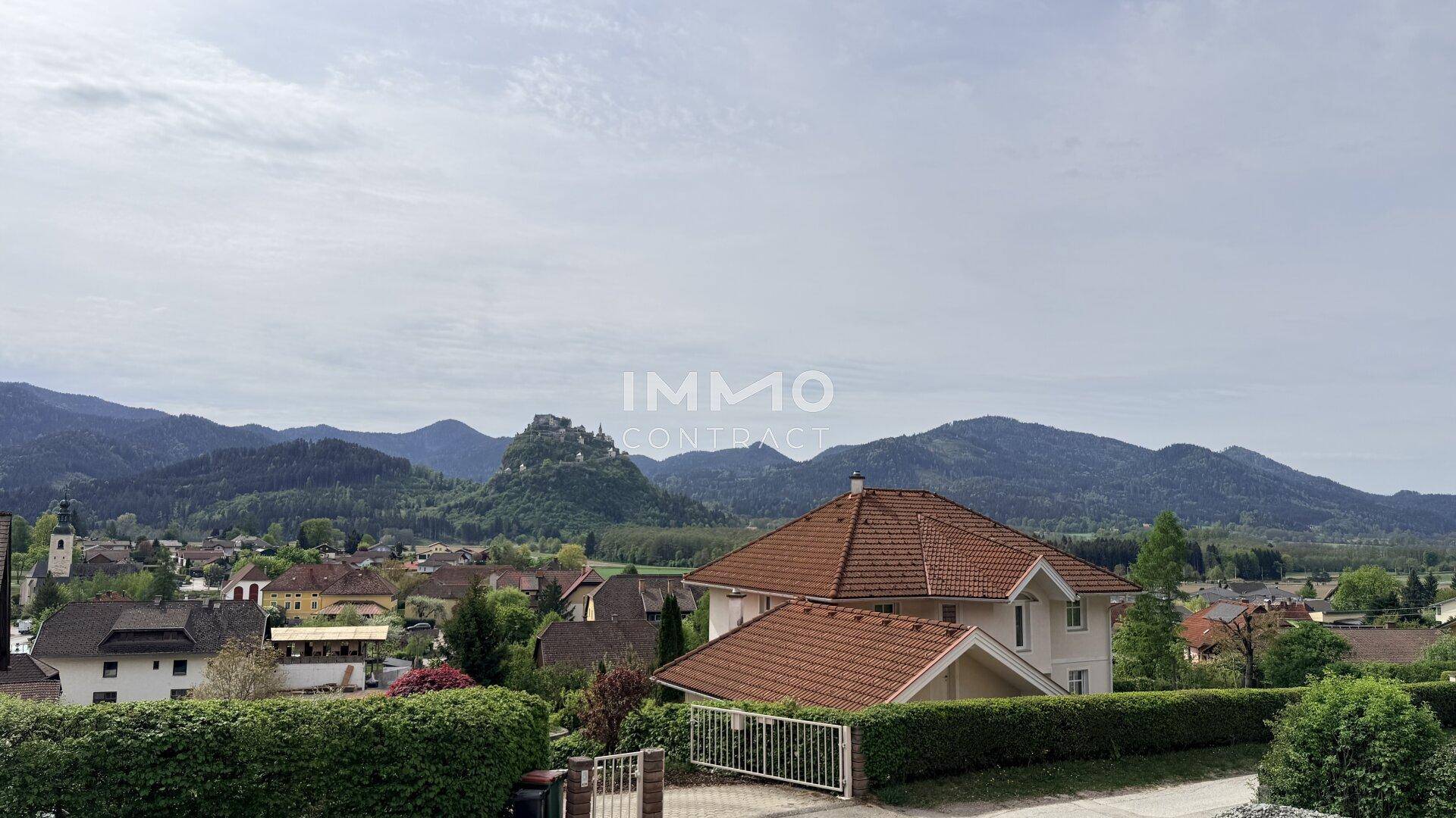 Panoramablick über eine malerische Ortschaft mit Bergen und einer Burg im Hintergrund.