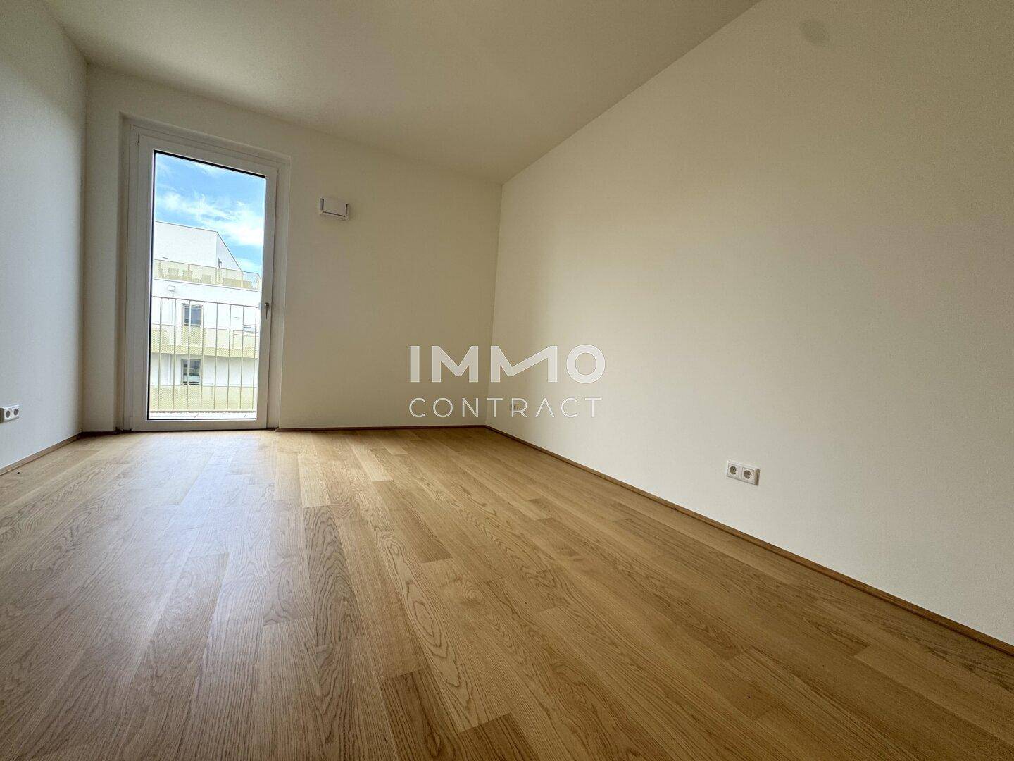 Leeres Zimmer mit Holzboden und einem Fenster, das Zugang zu einem französischen Balkon bietet.