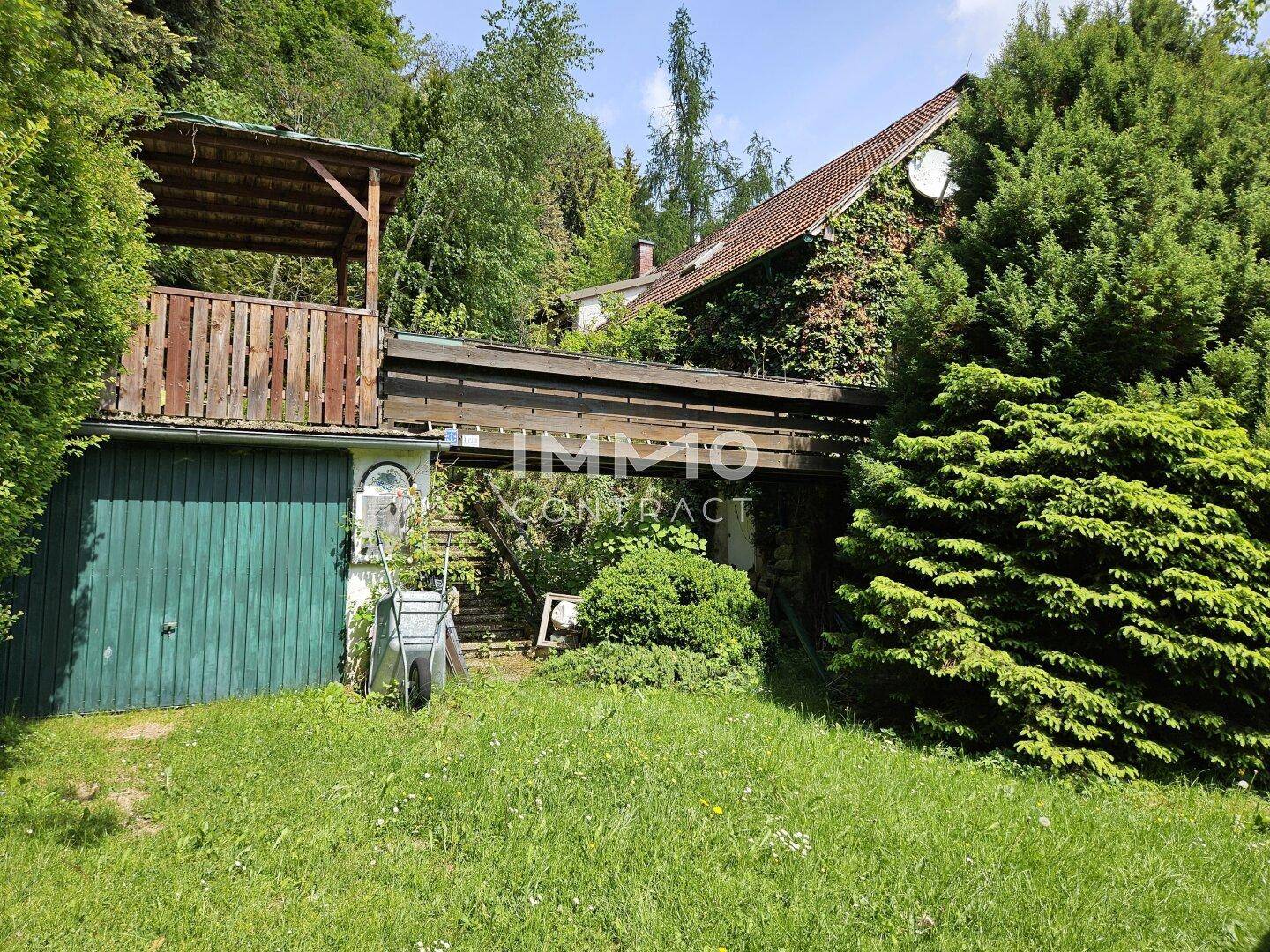 Rückseite des Hauses mit üppiger Vegetation, Garage und einer erhöhten Holzterrasse.