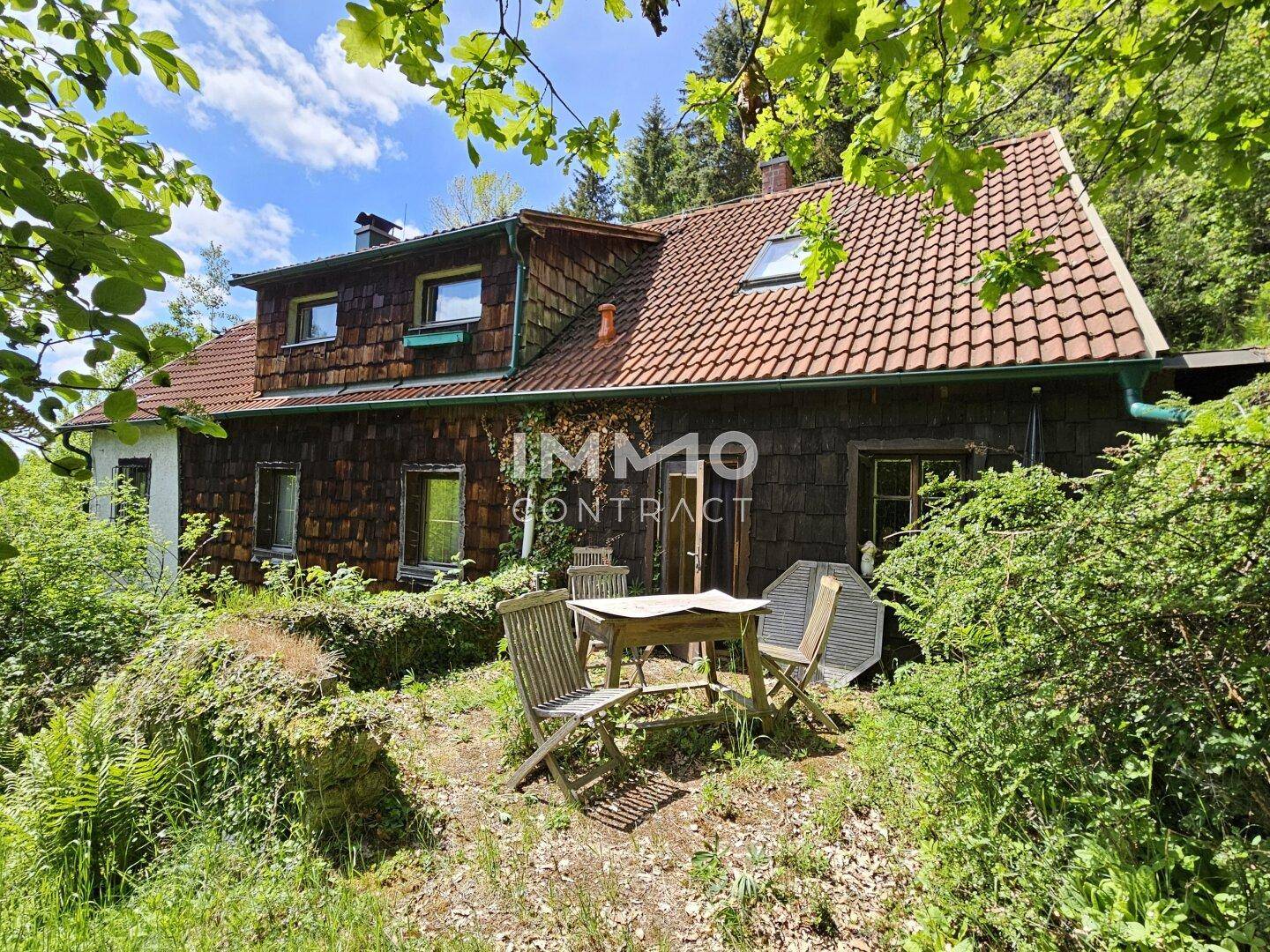 Rustikales Haus mit Holzschindeln und Ziegeldach, umgeben von einem naturbelassenen Garten.