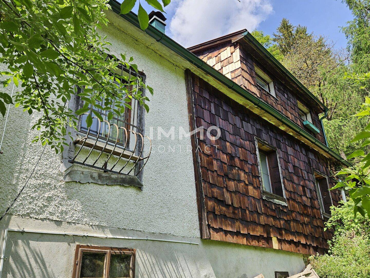 Seitenansicht des Hauses mit Putzfassade und Holzschindeln, umgeben von Bäumen und Sträuchern.
