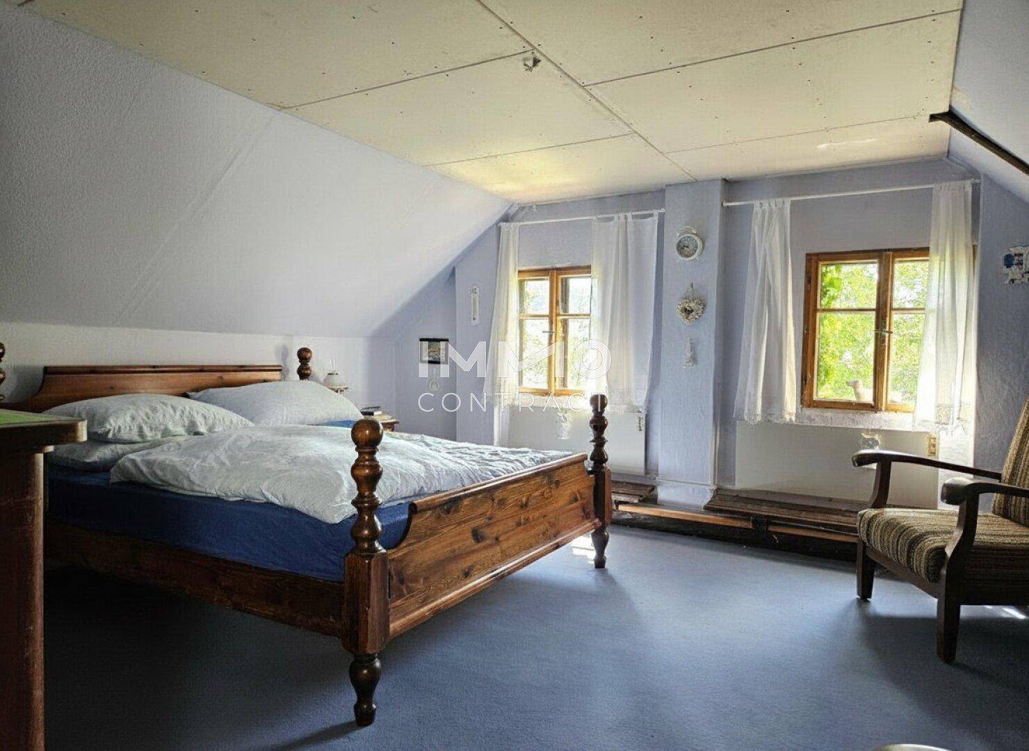 Schlafzimmer im Dachgeschoss mit Holzbett, blauer Auslegware und zwei Fenstern, hell und geräumig.