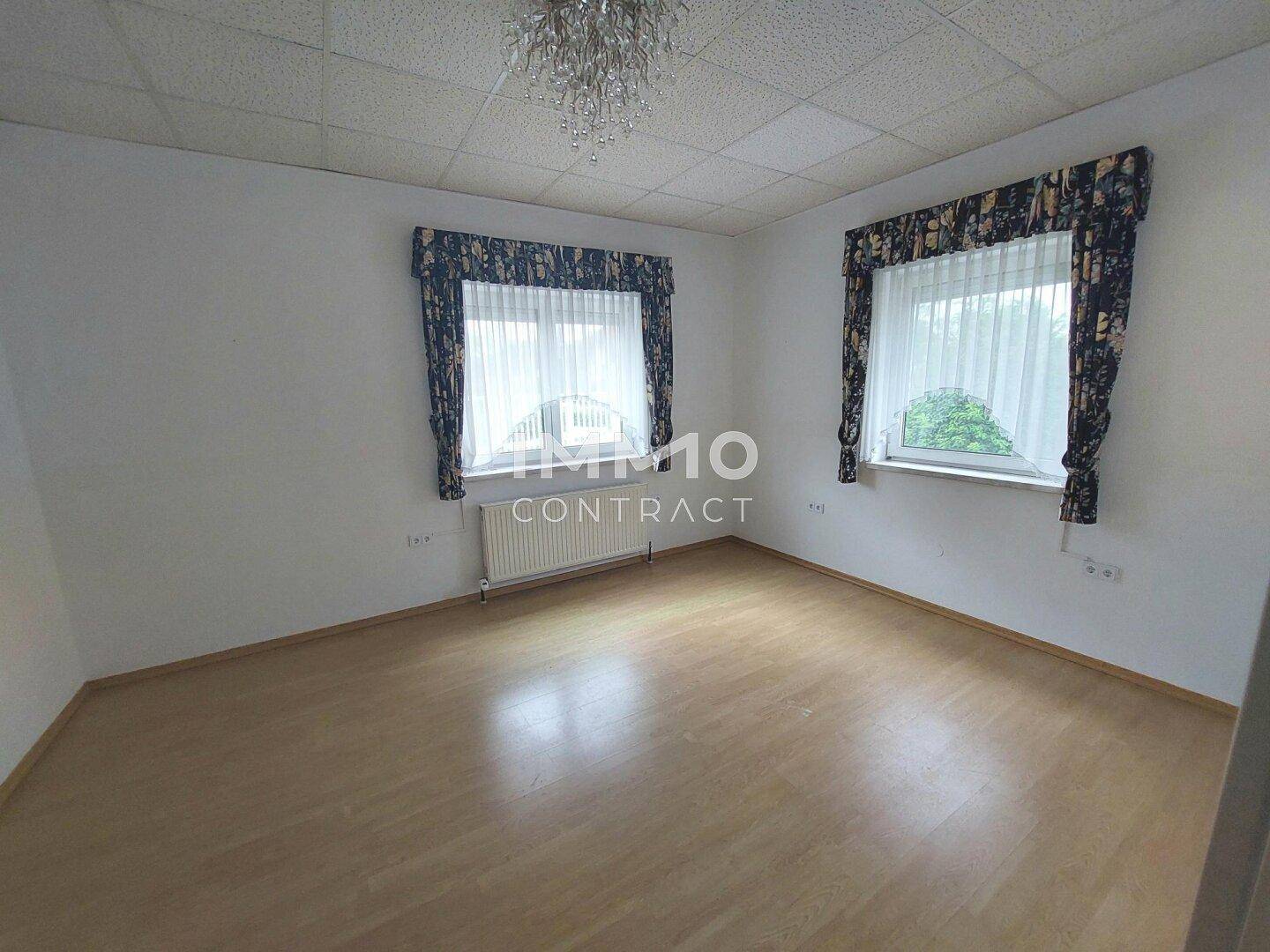 Helles Schlafzimmer mit Laminatboden, zwei Fenstern und einem Kronleuchter an der Decke.