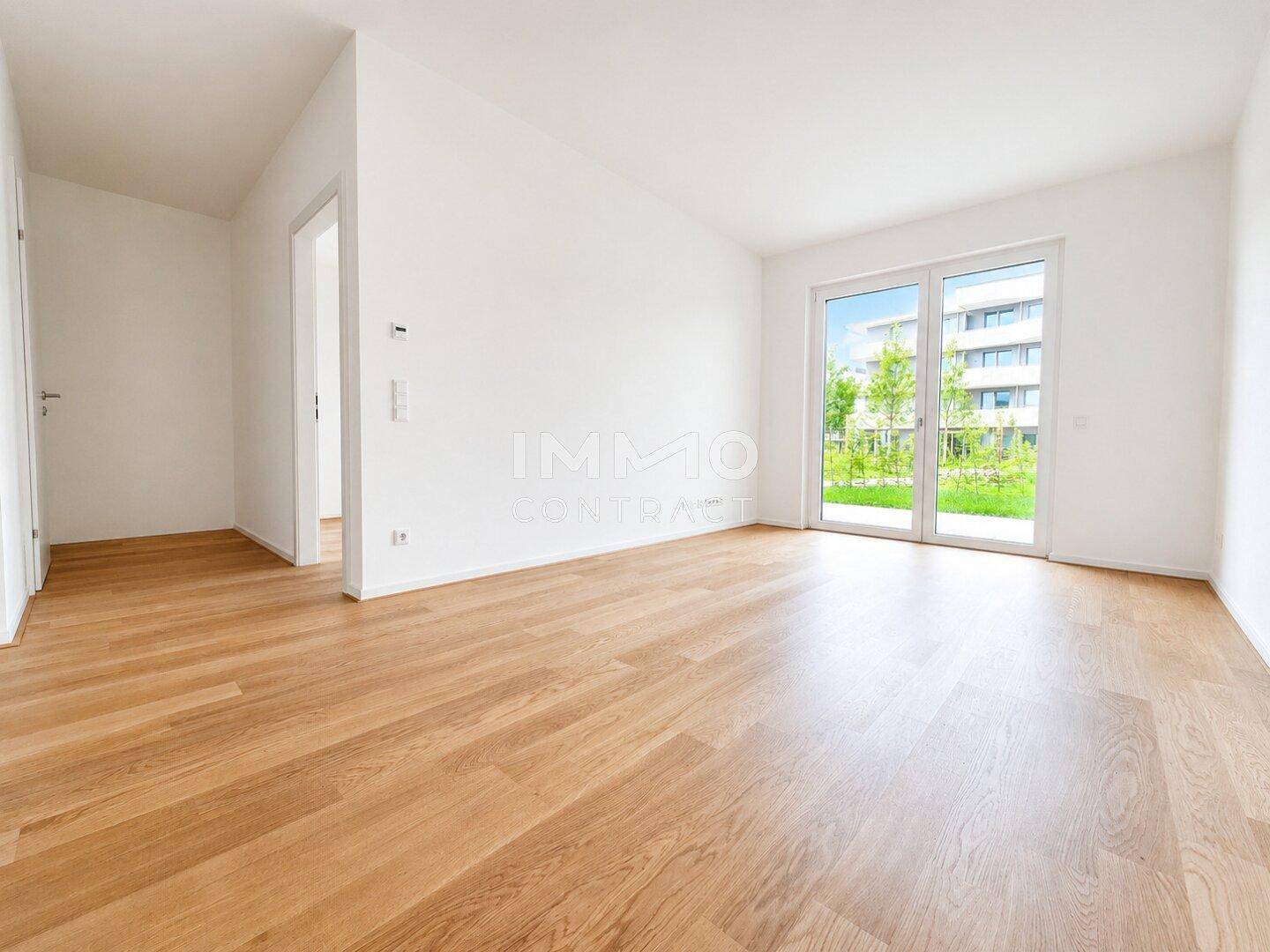 Heller, leerer Raum mit Holzboden und großen Fenstertüren, die zum Garten führen.