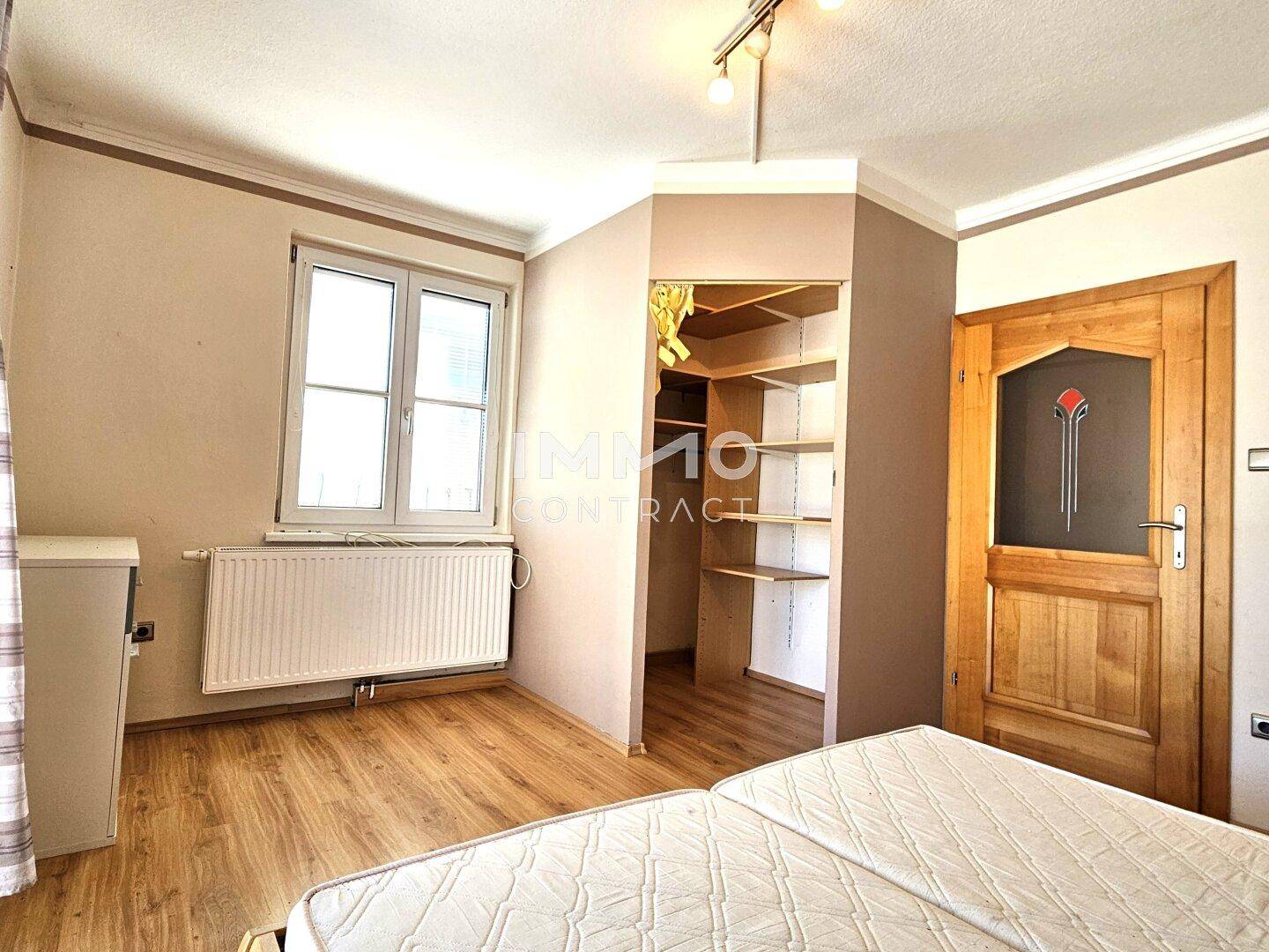 Schlafzimmer mit Laminatboden, Fenster und offenem Kleiderschrankbereich, hell und funktional.
