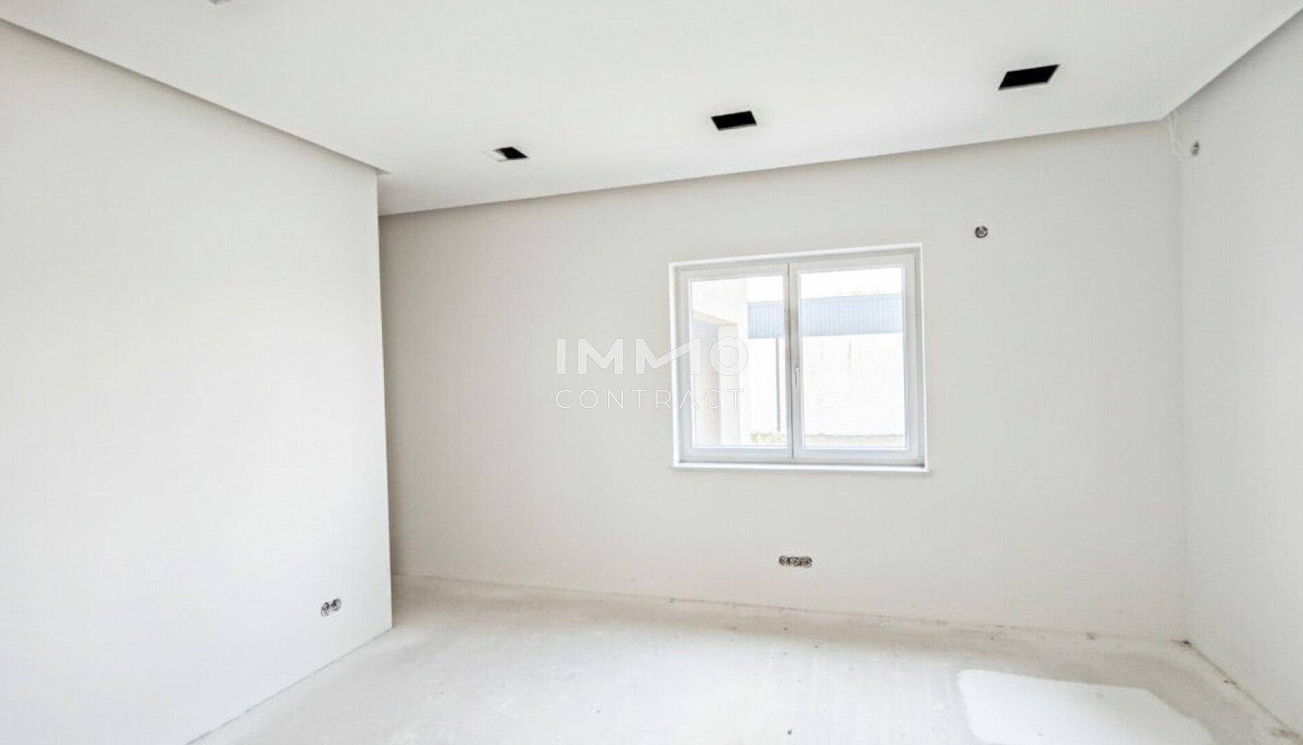 Kleiner Raum im Rohbau mit weißen Wänden, Betonboden und einem einfachen Fenster.