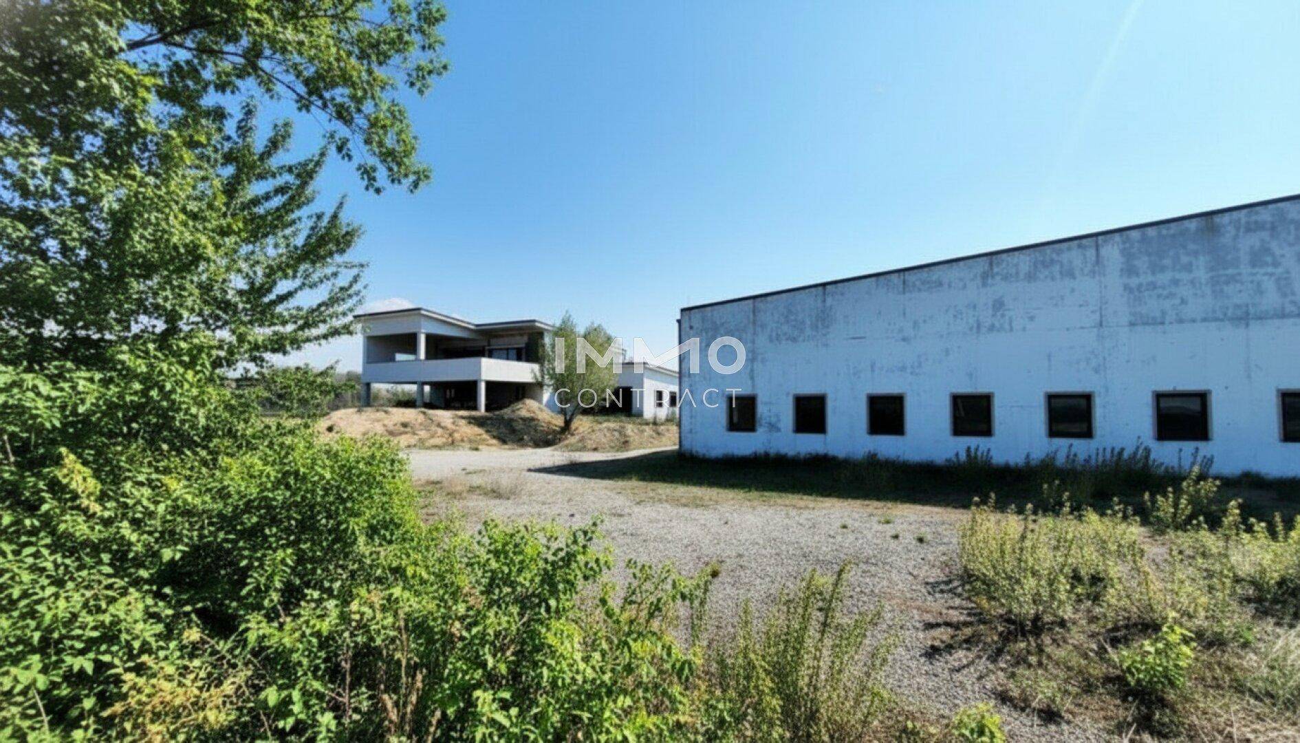 Außenansicht zweier Gebäude, eines modernen Rohbaus und einer großen Halle, umgeben von Vegetation.