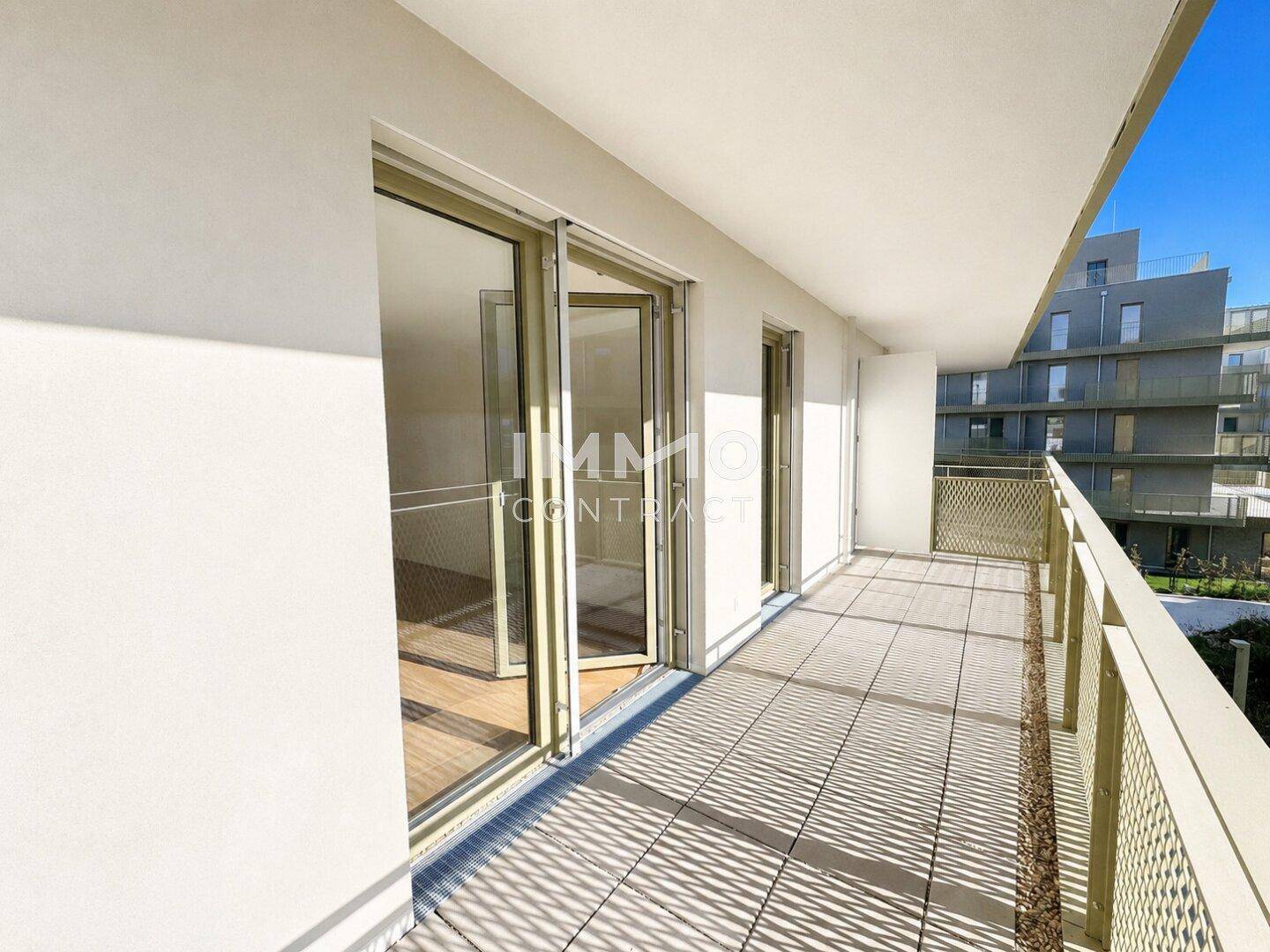 Langer Balkon mit Fliesenboden und Metallgeländer, bietet Blick auf moderne Gebäude.