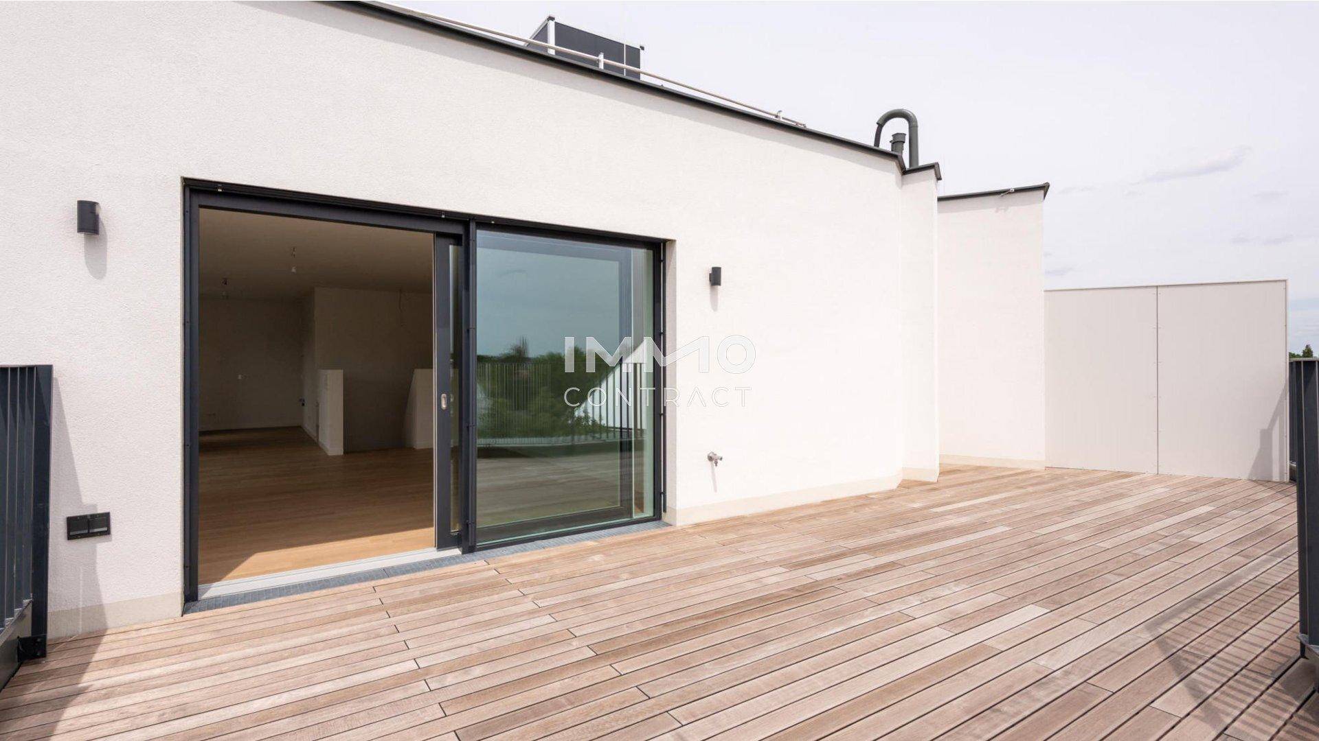 Große Holzterrasse mit Zugang zu einem hellen Innenraum durch eine Glasschiebetür.